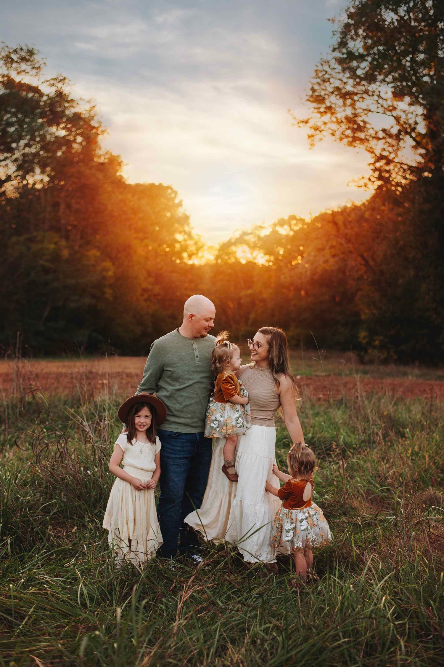 Family of five poses in golden meadow at sunset, parents holding toddler while two daughters stand nearby.