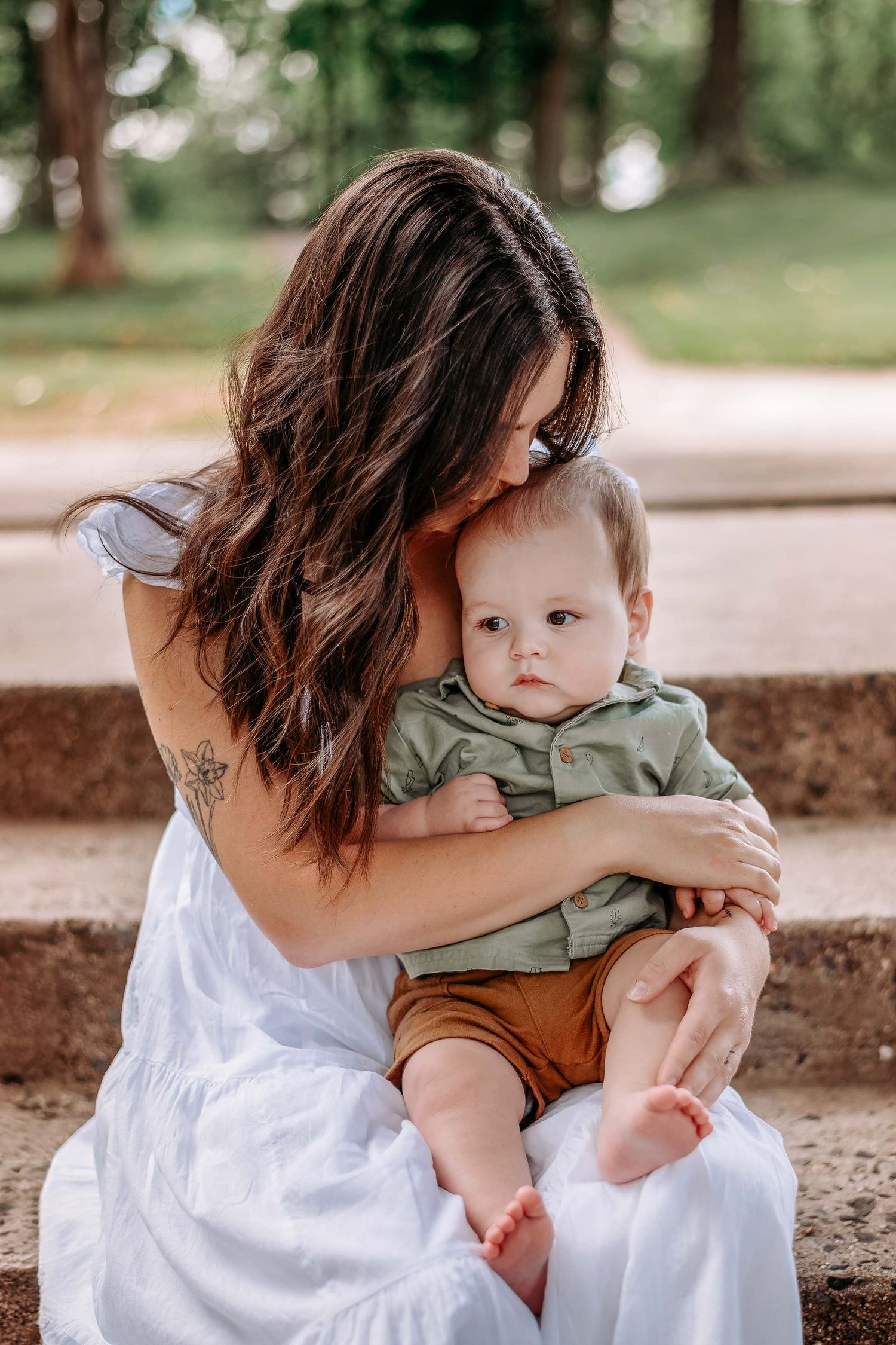 A young mother with long brown hair in a white dress tenderly kisses her baby boy outdoors.