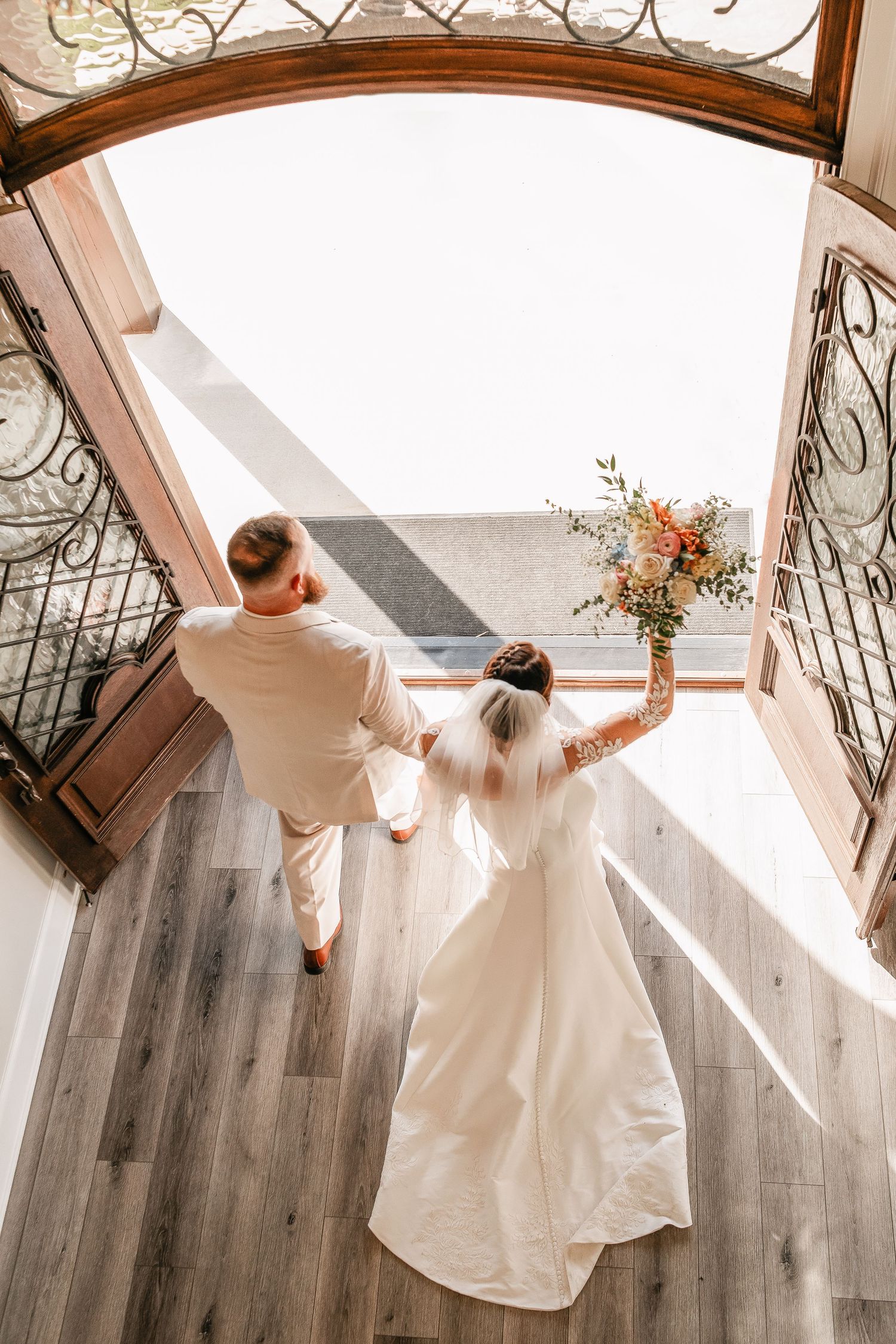 Aerial view of bride in white gown and groom in tan suit holding hands, walking through ornate arched doors.