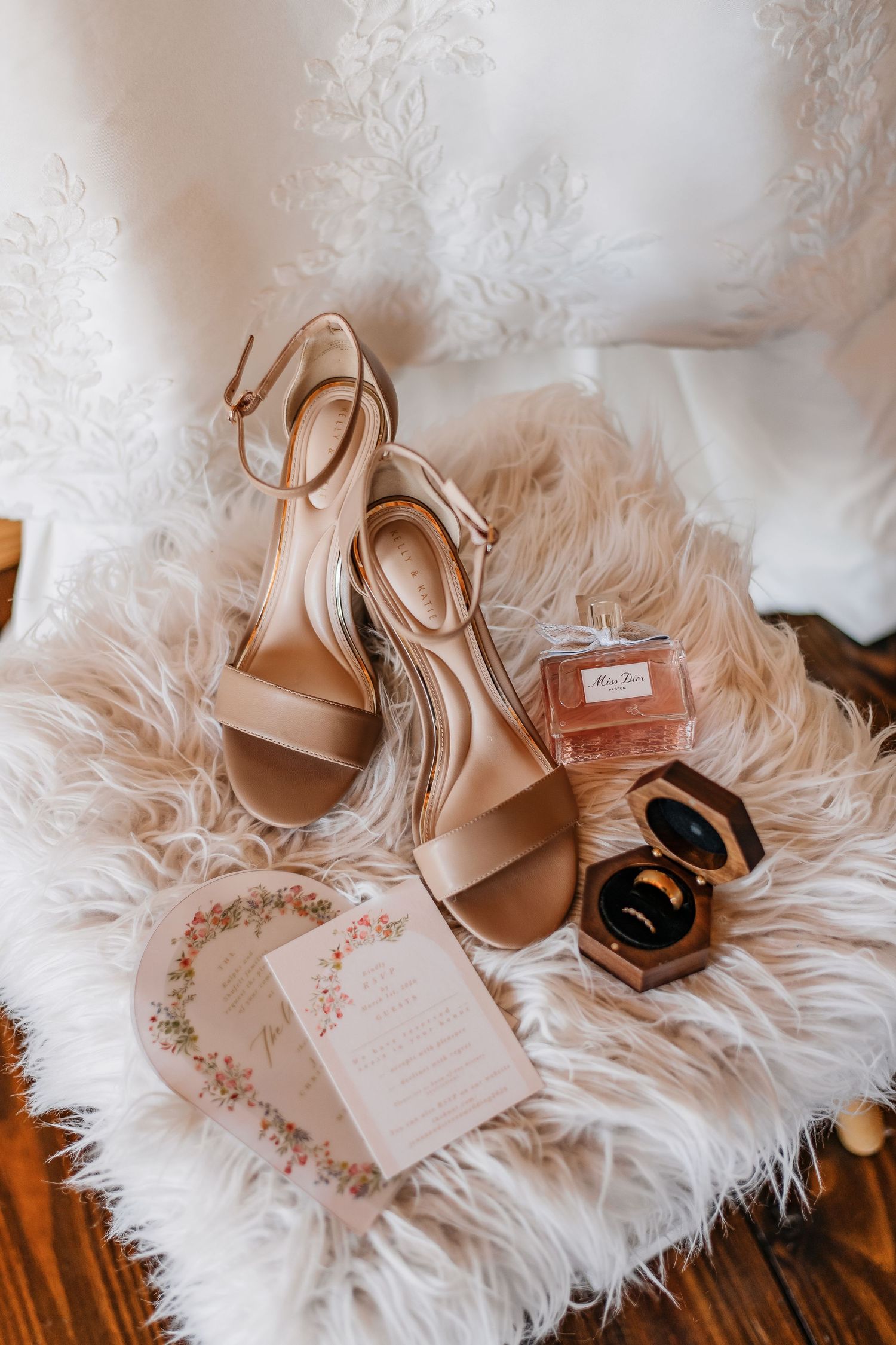 Bridal flat lay featuring nude heels, Miss Dior perfume, gold rings in velvet box, and floral invitation on fur.