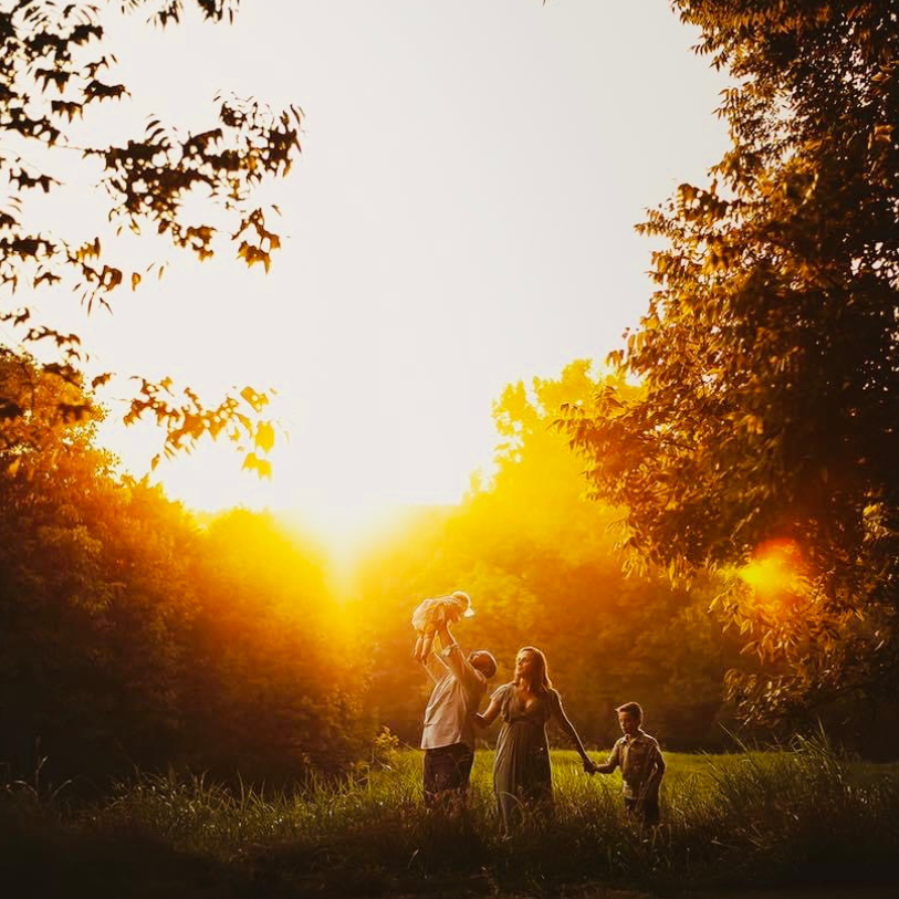 A silhouetted family walks together during golden sunset through a grassy field with trees in the background.