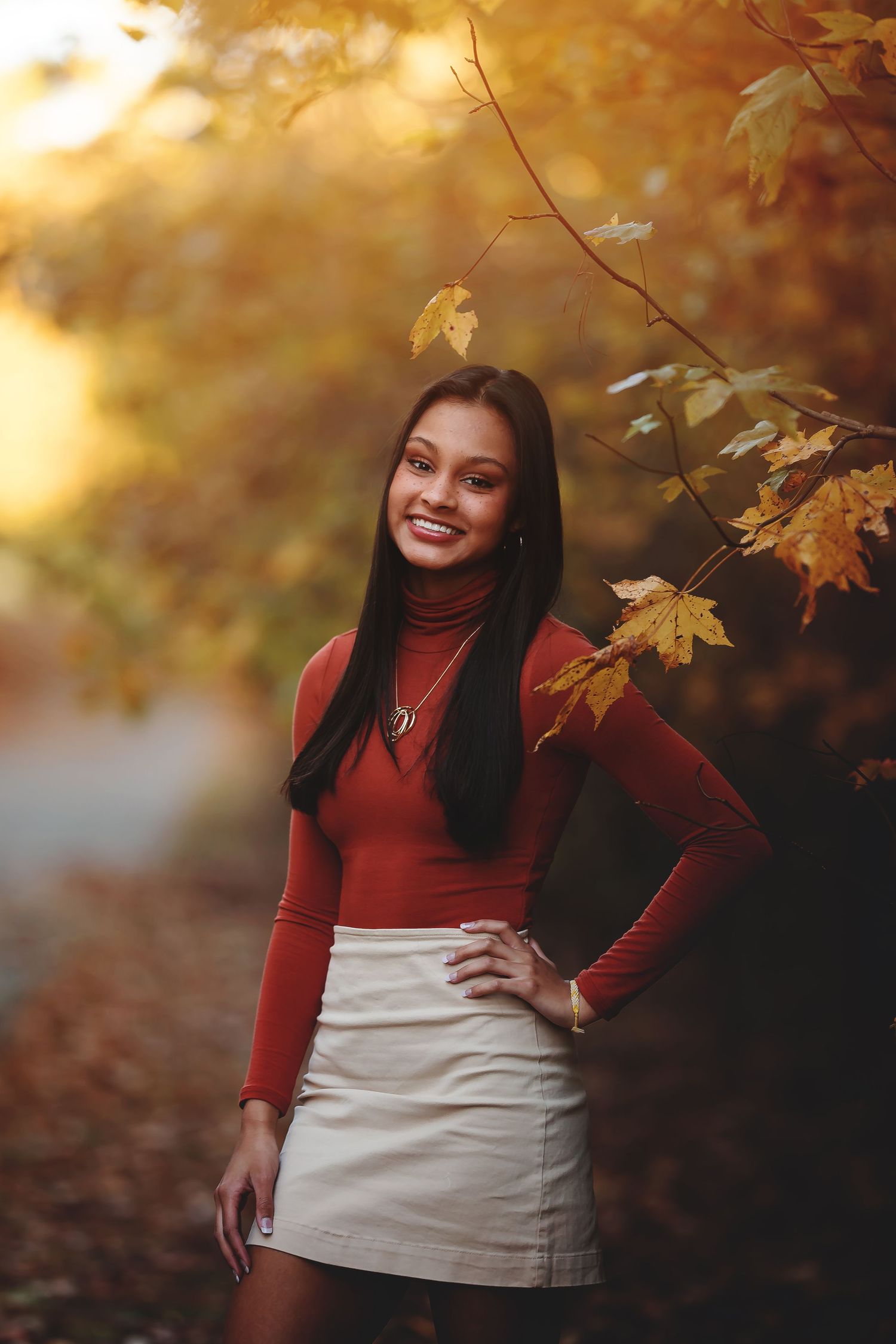 Woman in red sweater and white skirt smiles under golden autumn leaves.