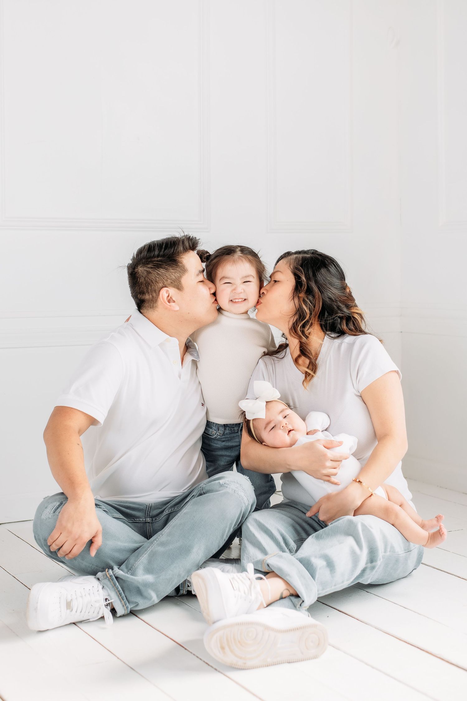Family sitting together on white floor wearing casual clothing in bright minimal studio setting.