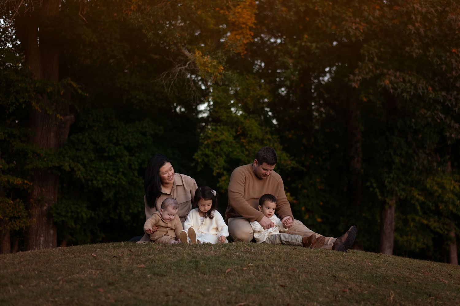 Family group photo session at sunset on a grassy hill surrounded by trees.
