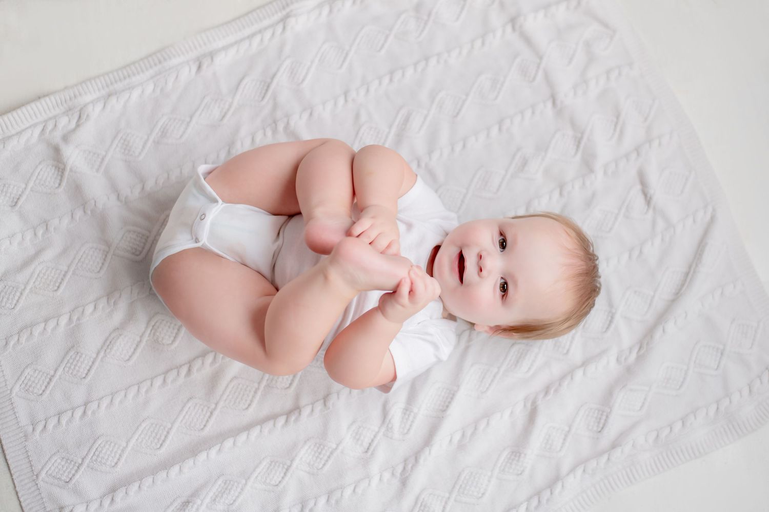 A baby in a white diaper lies on a textured white blanket while smiling and reaching for their toes.
