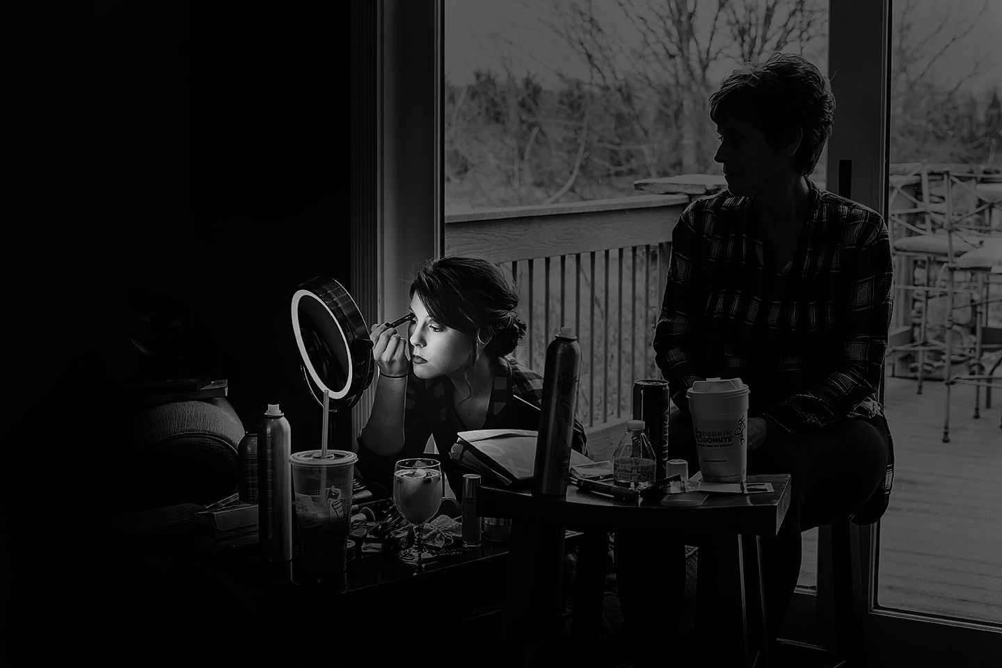 Black and white photo of a woman applying makeup at a vanity mirror while another person stands nearby by a window.