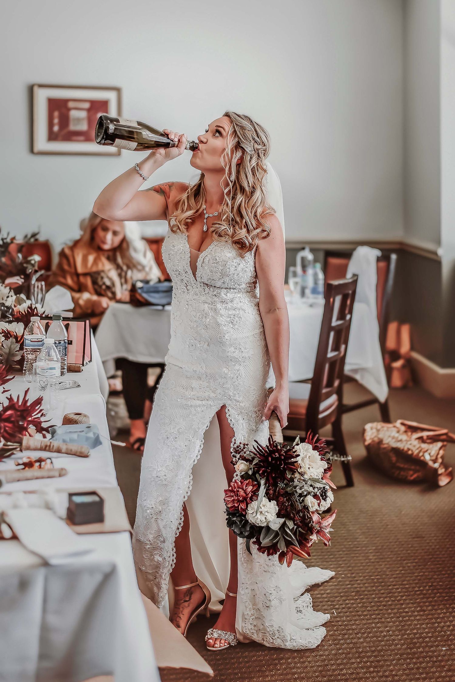 Bride in lace wedding gown drinking from champagne bottle while holding bouquet at wedding reception.