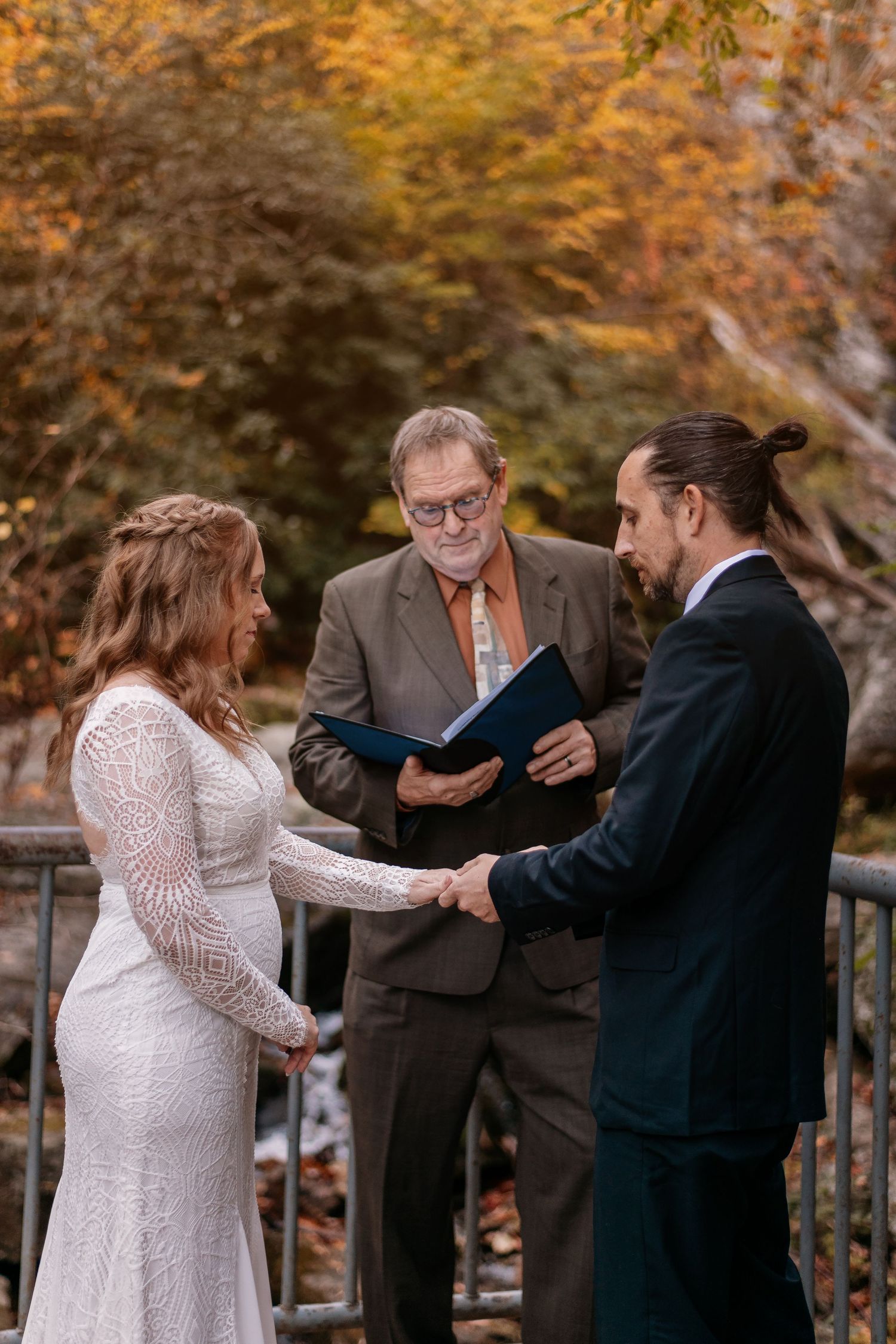 Couple exchanging vows outdoors in fall with officiant holding blue folder during intimate wedding ceremony.