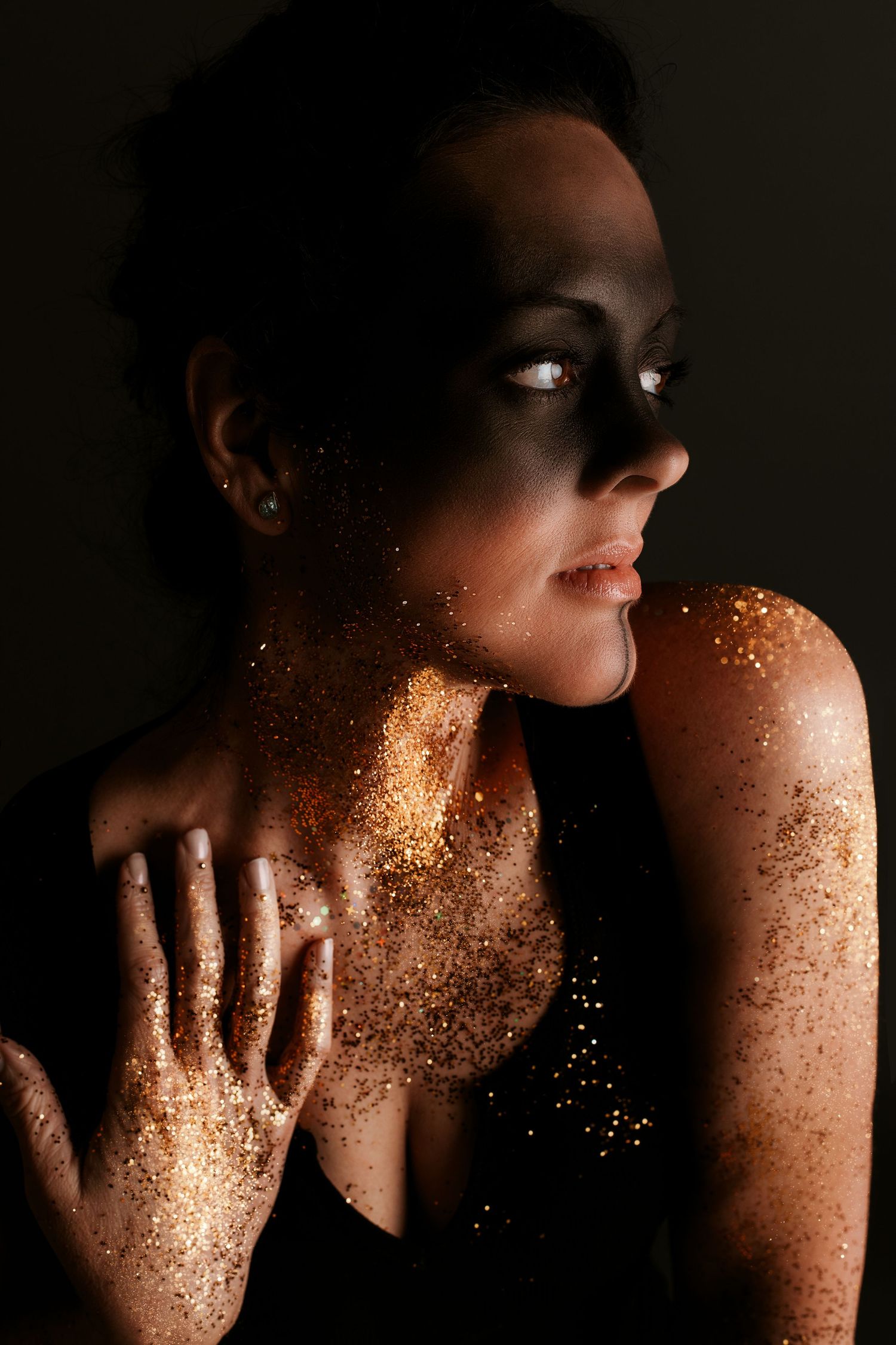 A dark artistic portrait with gold glitter sparkling on skin against a black background in dramatic lighting.