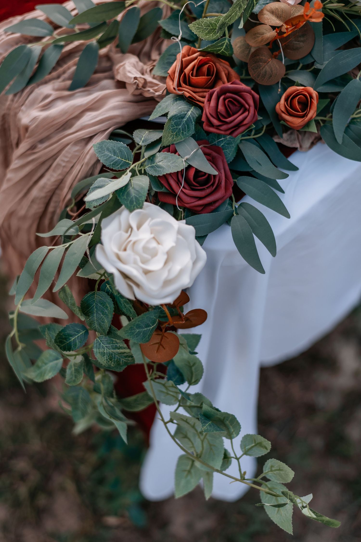 Elegant wedding floral arrangement with white, burgundy, and terracotta roses with eucalyptus greenery on a white tablecloth.