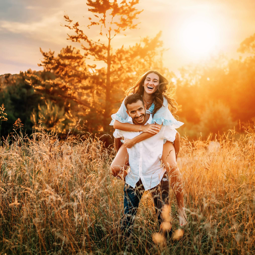 Couple playfully running through a golden field of tall grass at sunset with warm orange lighting and pine trees.