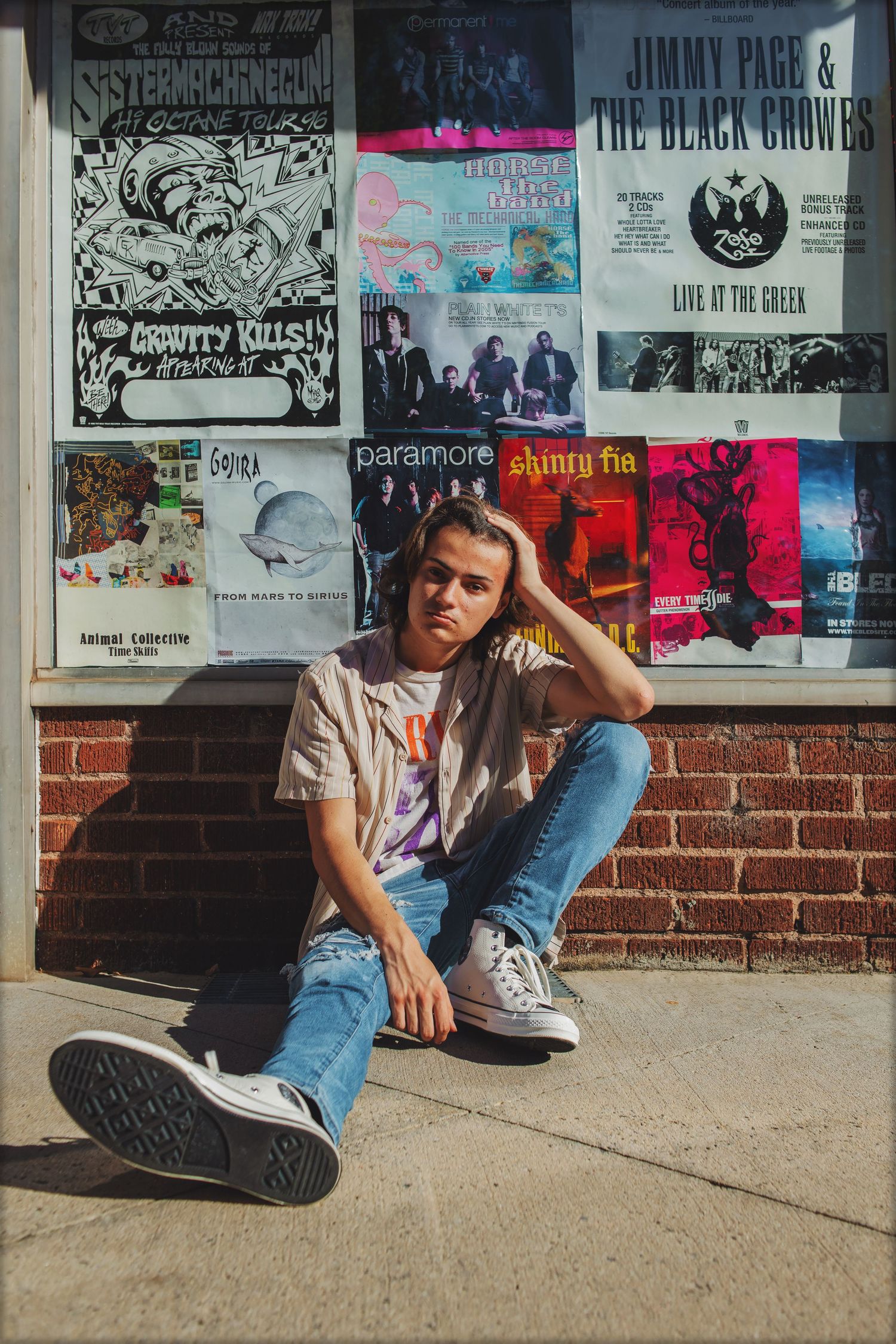 Person in casual attire sitting against brick wall with vintage music posters displayed above.
