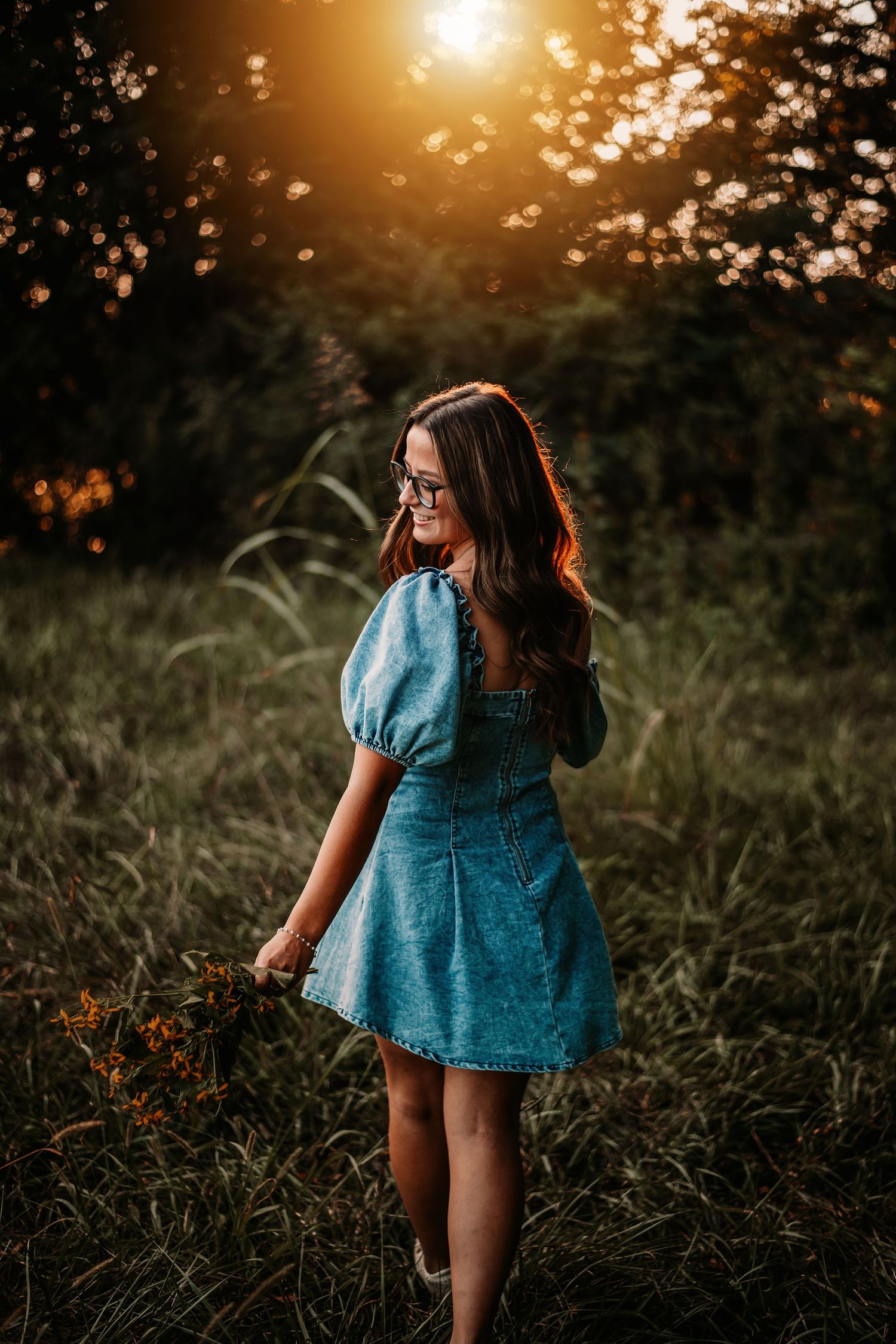 Woman in blue dress walking through tall grass at sunset.