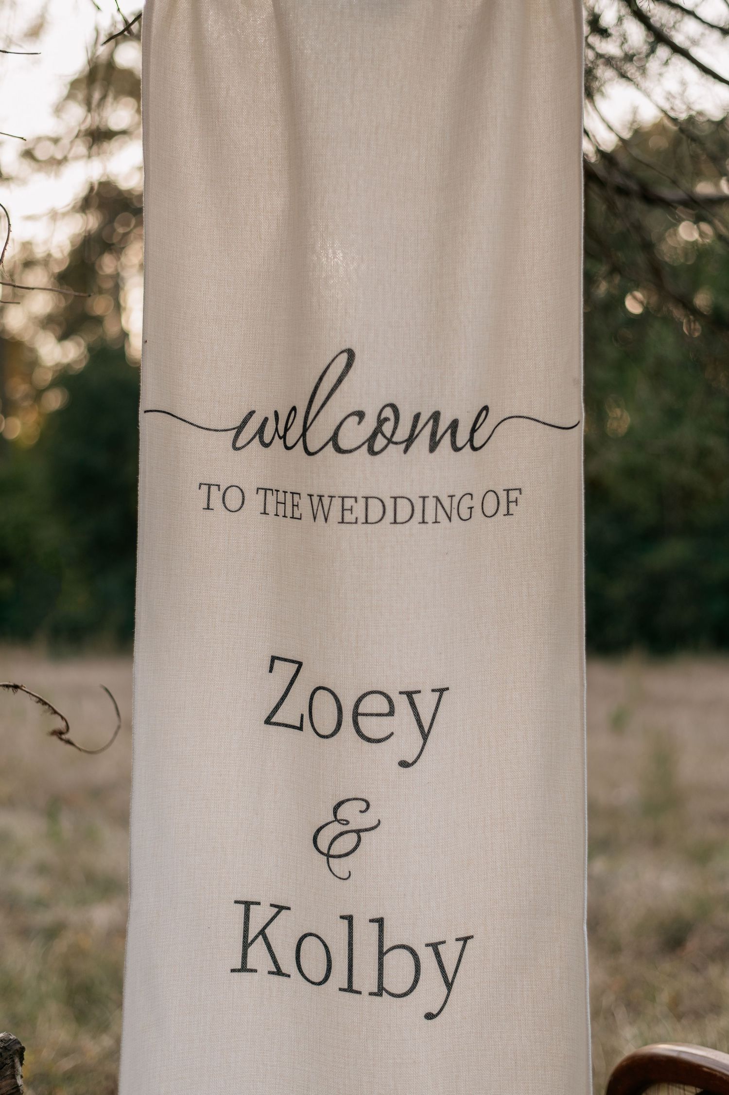 Elegant outdoor wedding welcome banner for Zoey and Kolby, displayed on cream fabric among trees.