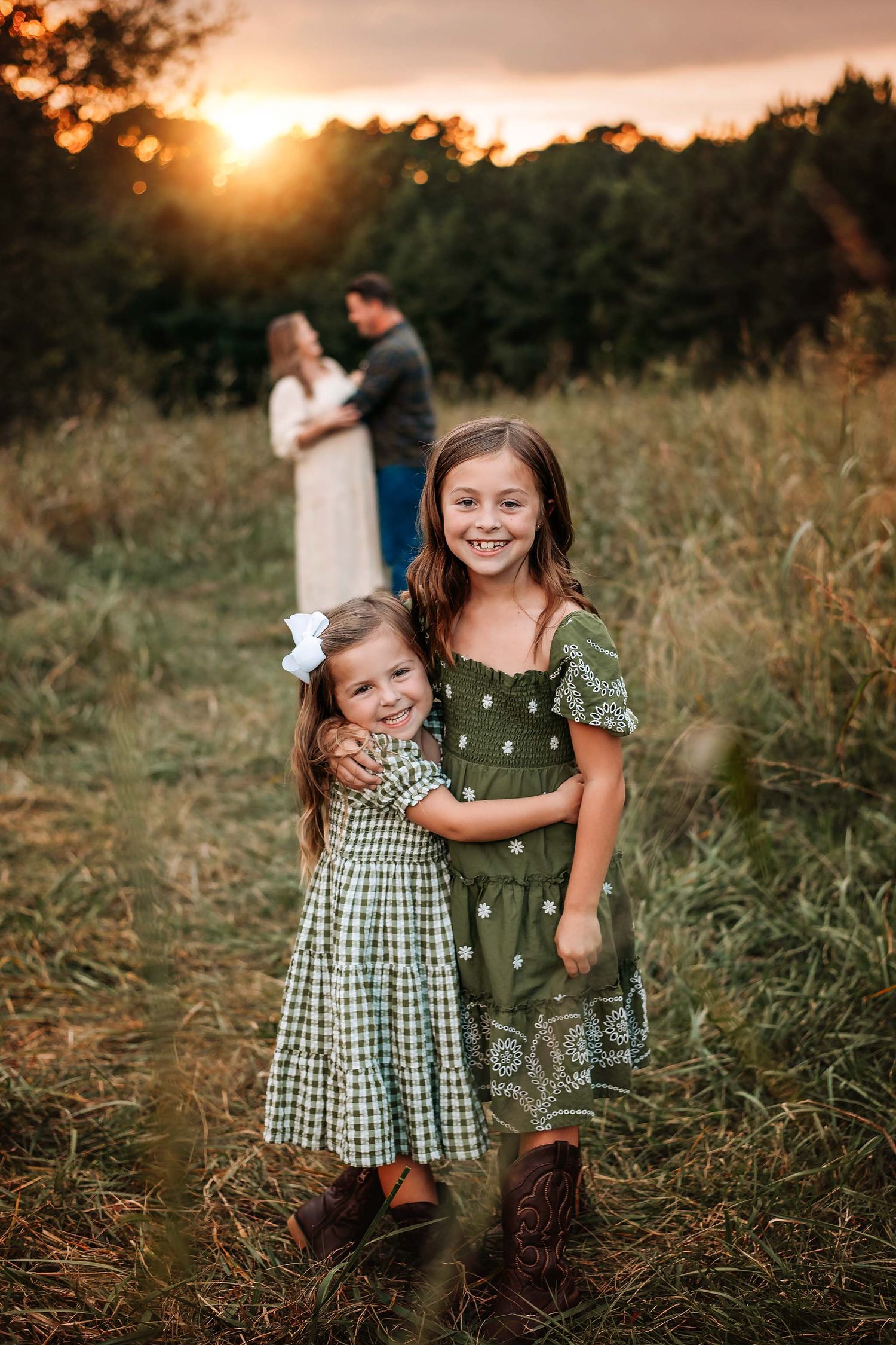 Two young sisters in green dresses embrace in a field at golden sunset, with their parents standing together in the background.