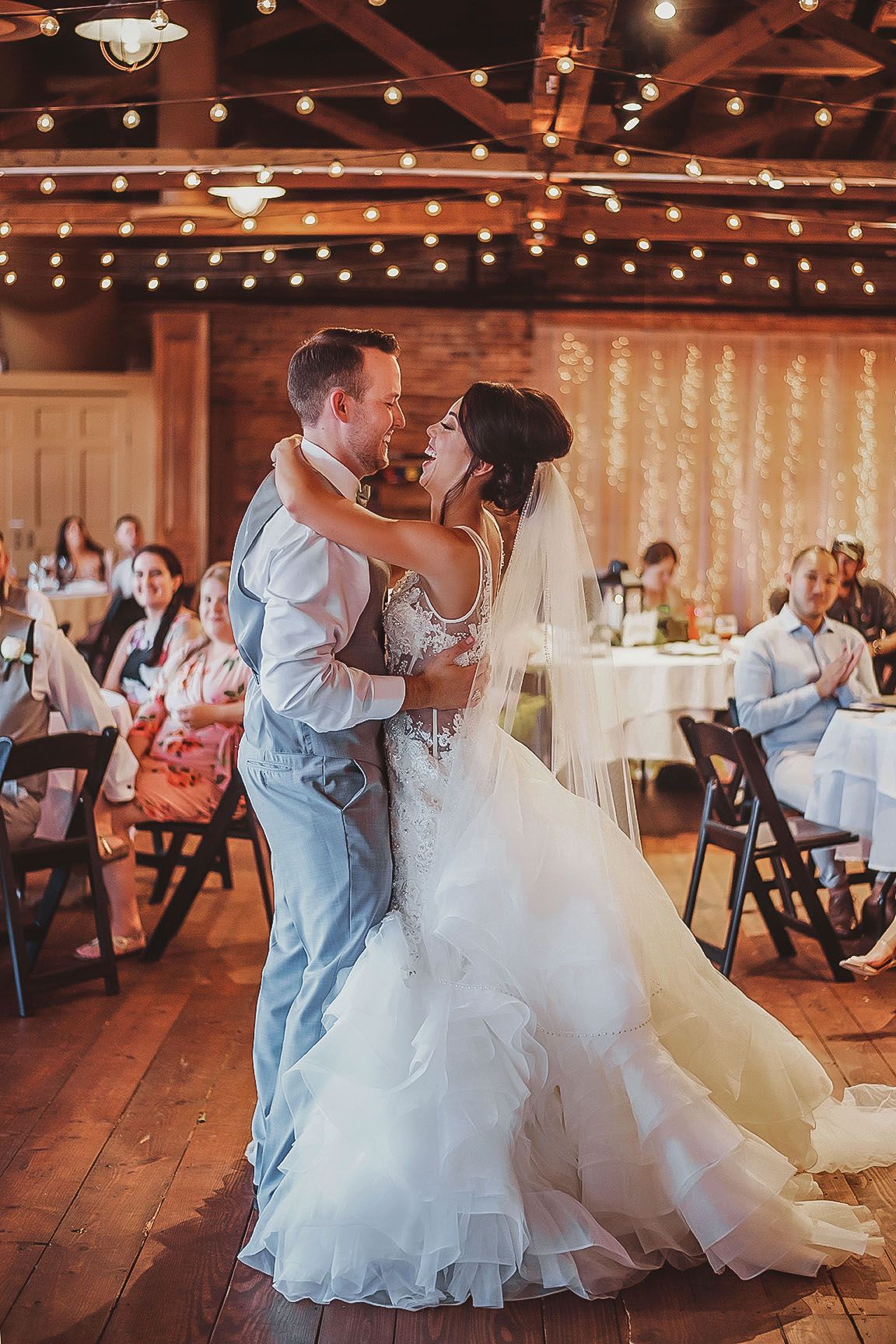 Bride and groom share their first dance in a warmly lit rustic wedding venue with string lights overhead.