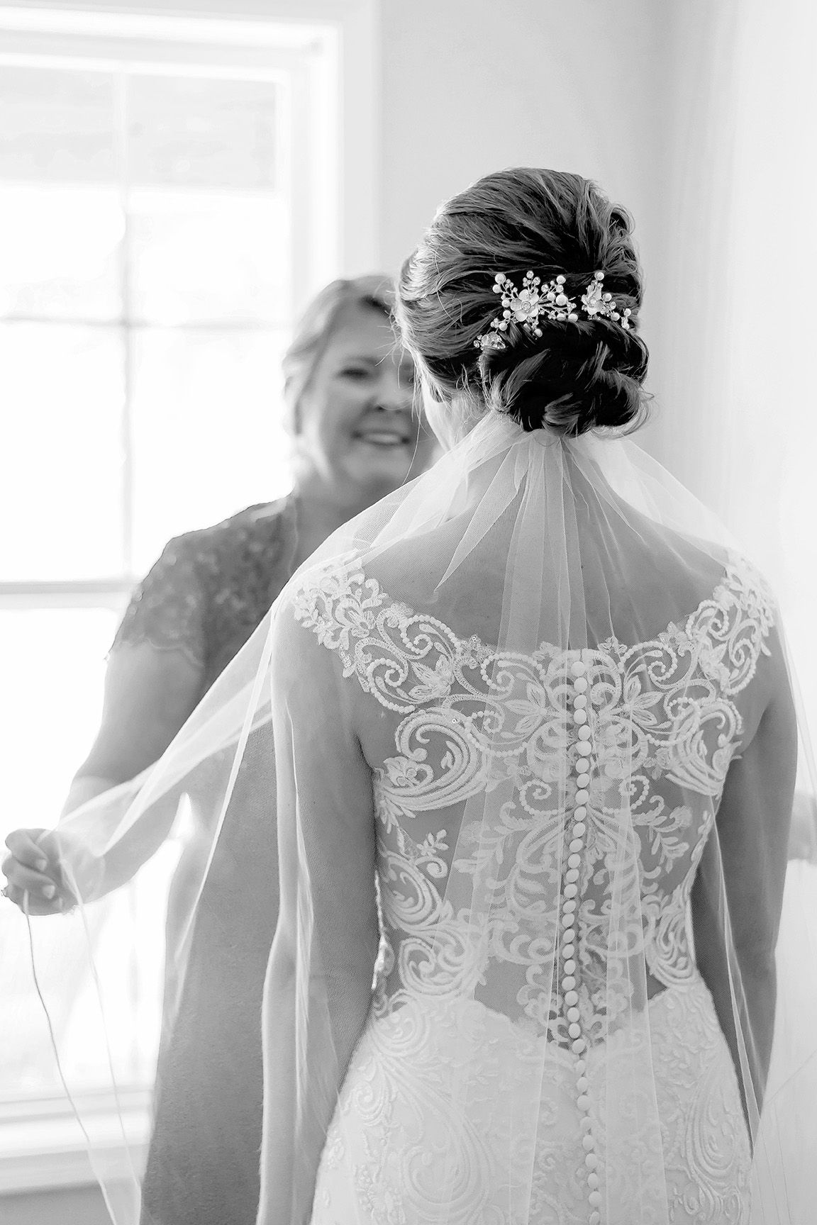 Black and white photo of bride in lace wedding dress with veil, showing intricate back detail, as woman adjusts her veil.