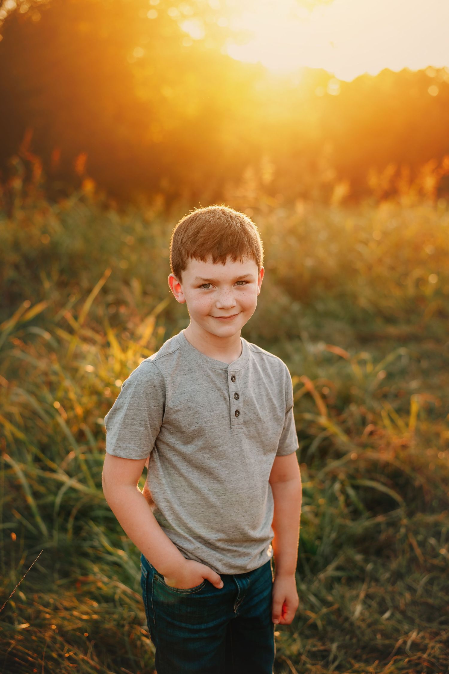 Young person in grey henley shirt standing in sunlit field at sunset.