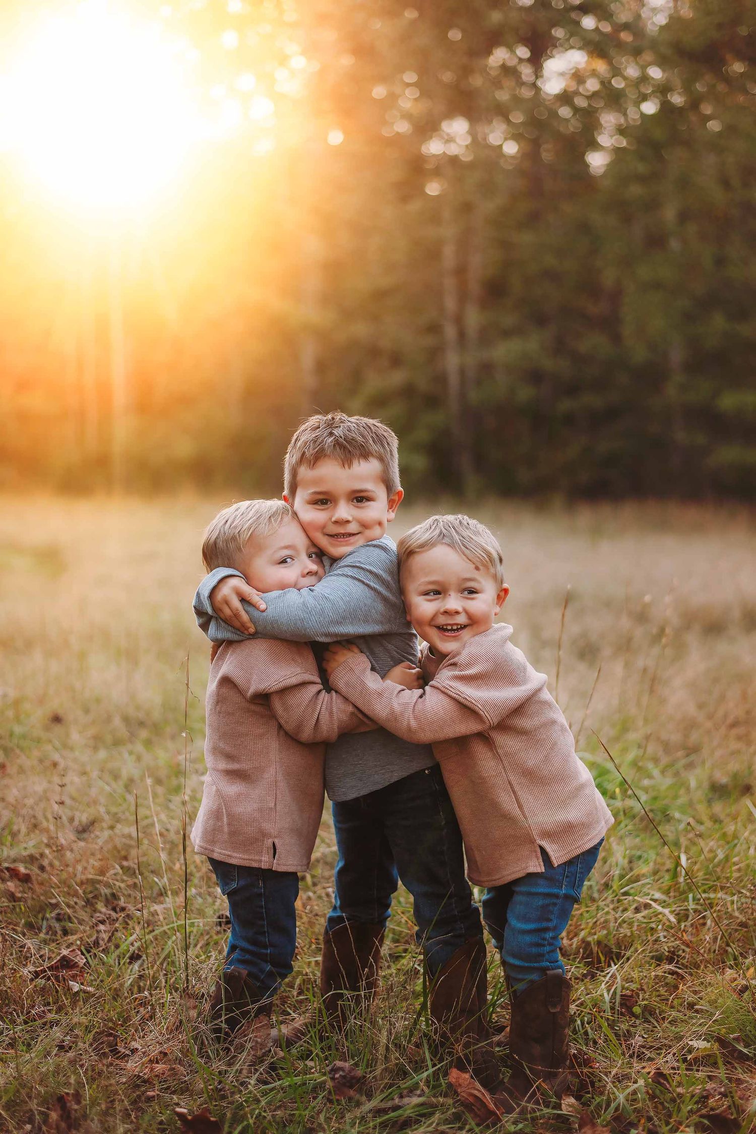 Three young children hugging each other in a golden sunset field, wearing matching coats, smiling outdoors.