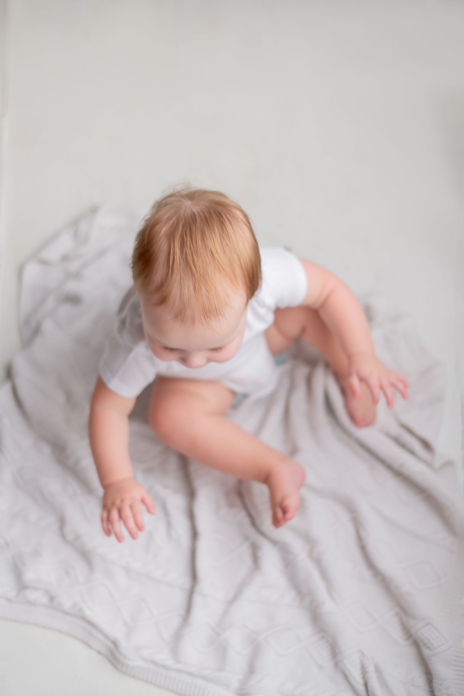 A baby in a white onesie sits on a soft gray blanket, viewed from above.