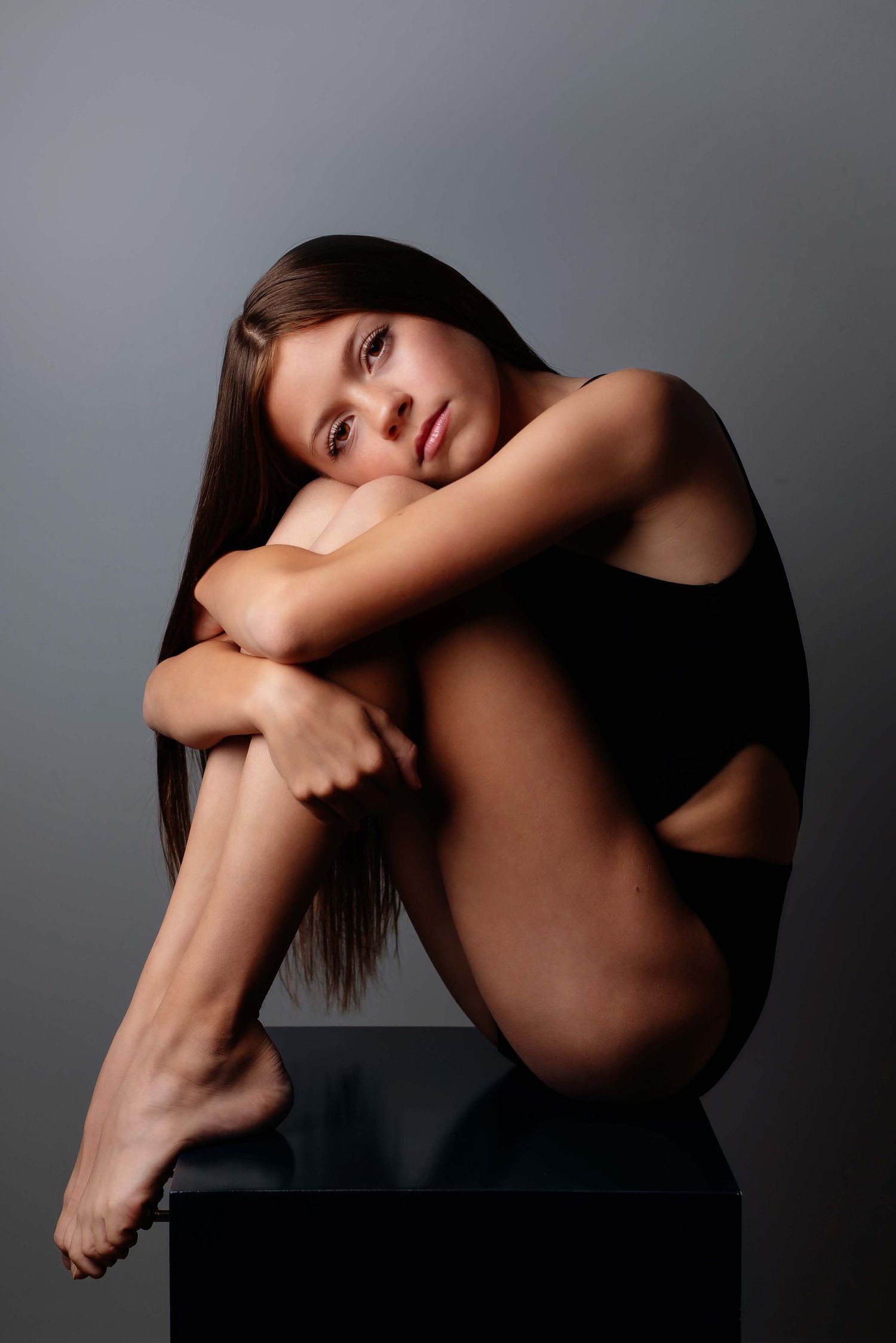 Young woman in black bodysuit sitting on a dark cube, hugging her knees, with long brown hair on gray background.