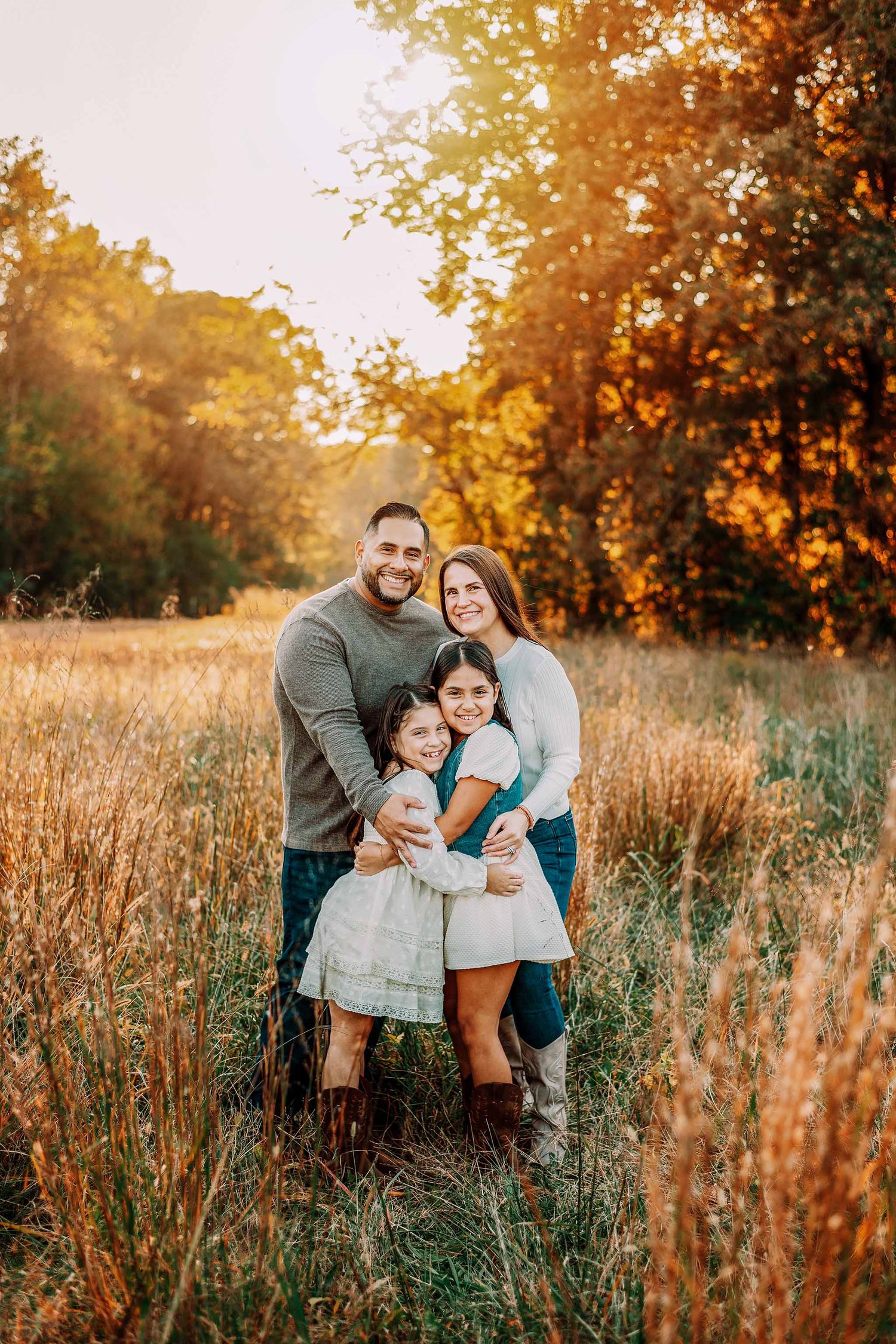 Family of four smiling together in a golden autumn field at sunset, parents hugging two young daughters.