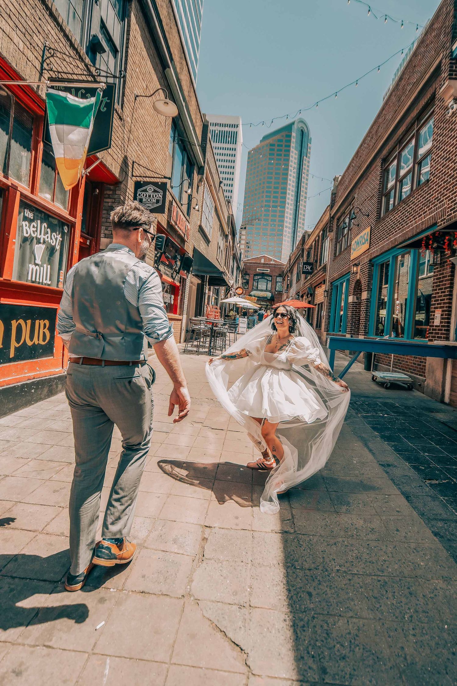 A bride in a white dress dances joyfully with her groom on a sunny urban street lined with colorful shops and skyscrapers.