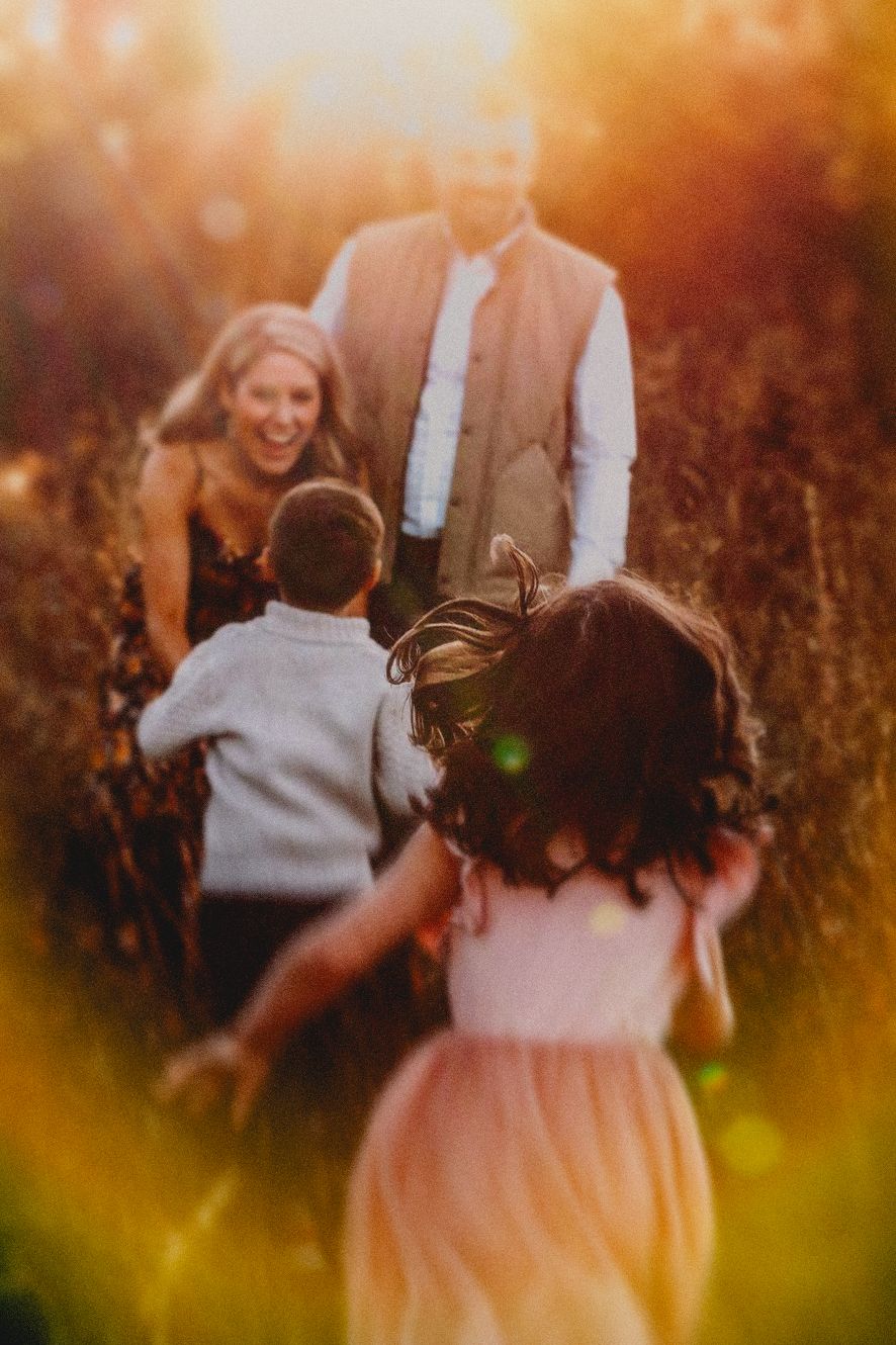 Family laughing together in golden sunlit field, children running toward parents in warm autumn outdoor portrait.