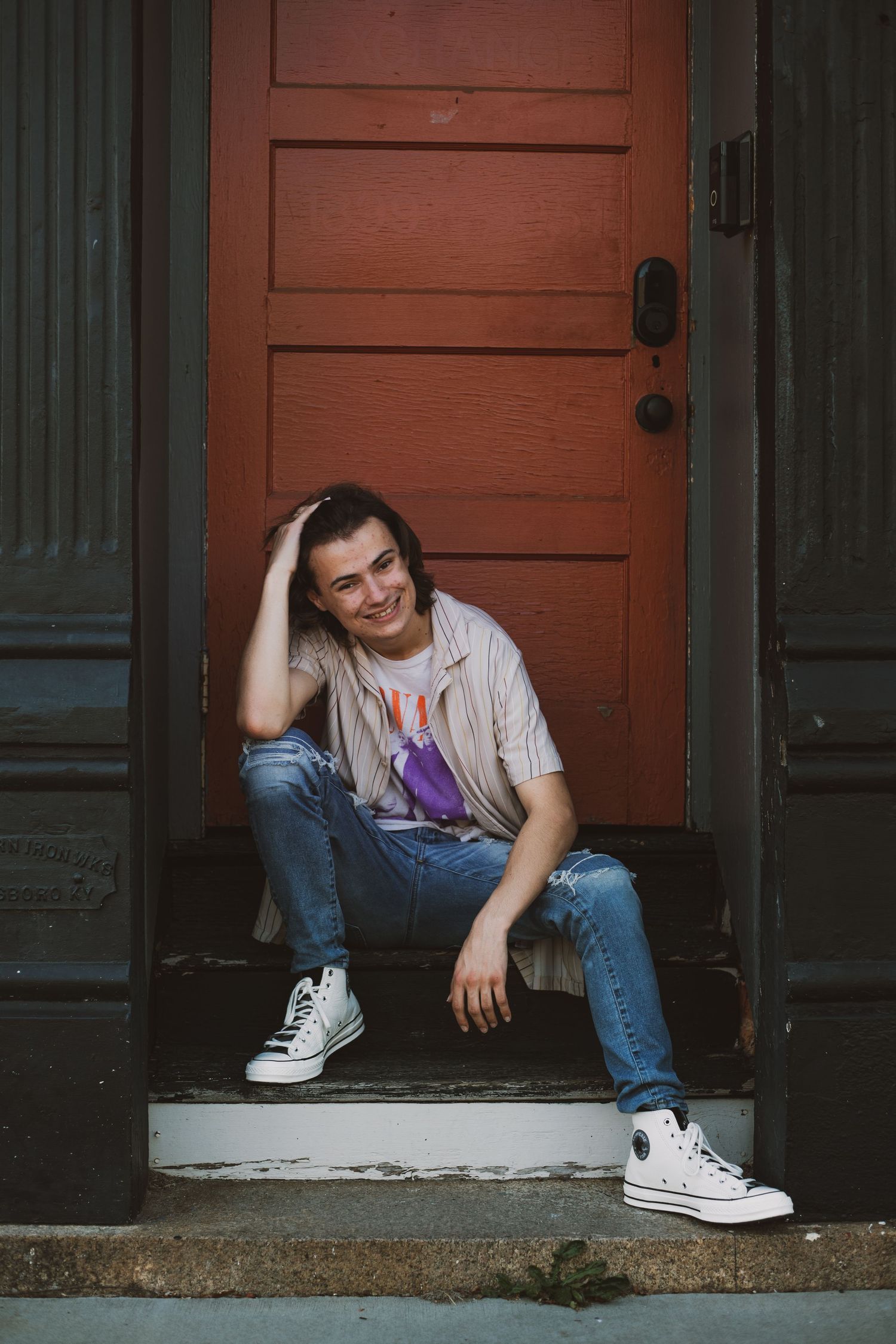 Person in casual wear sitting on steps in front of rustic red door.
