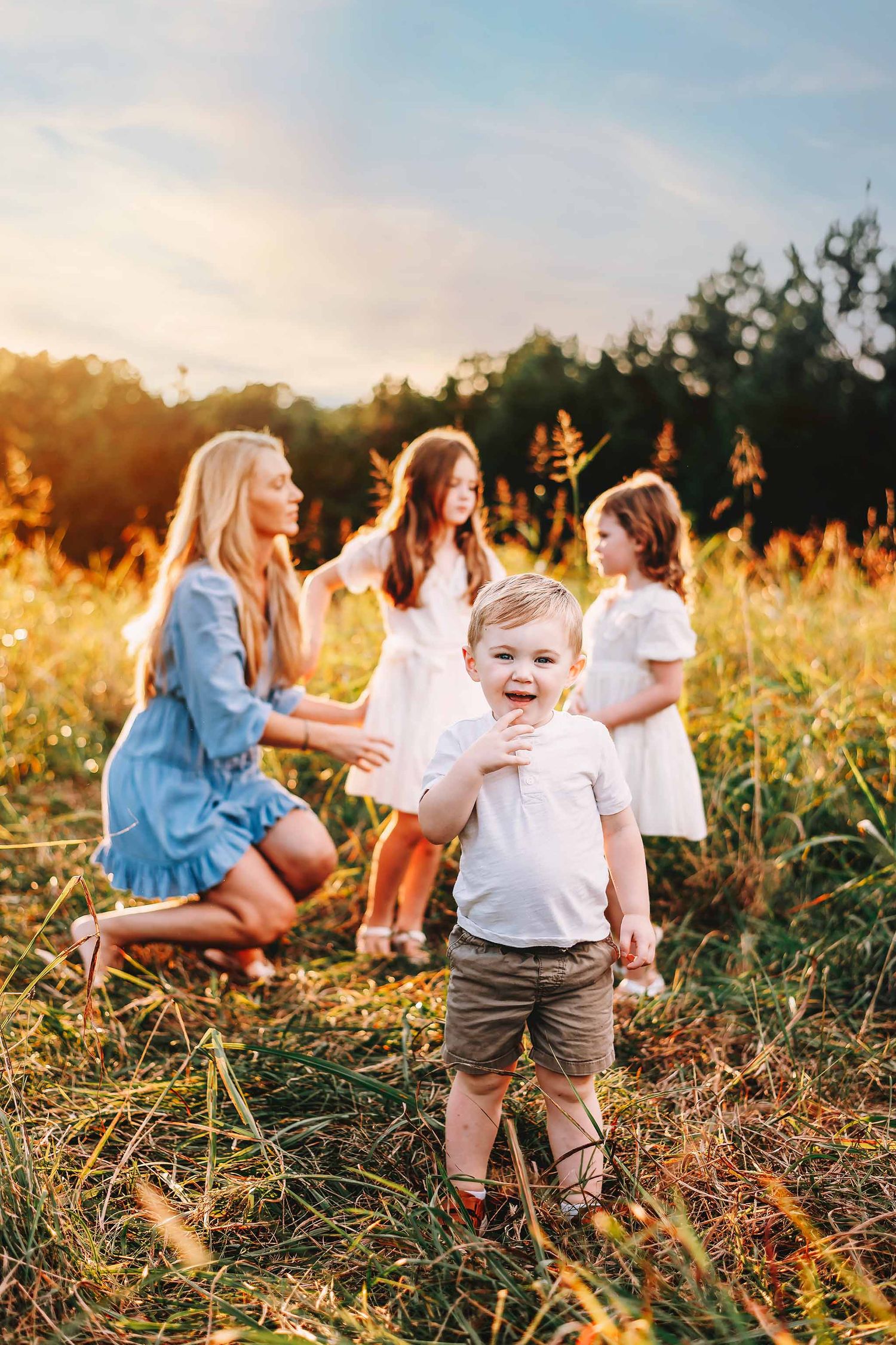 A joyful toddler boy stands in a sunny field while a woman and two girls play in the golden light behind him.