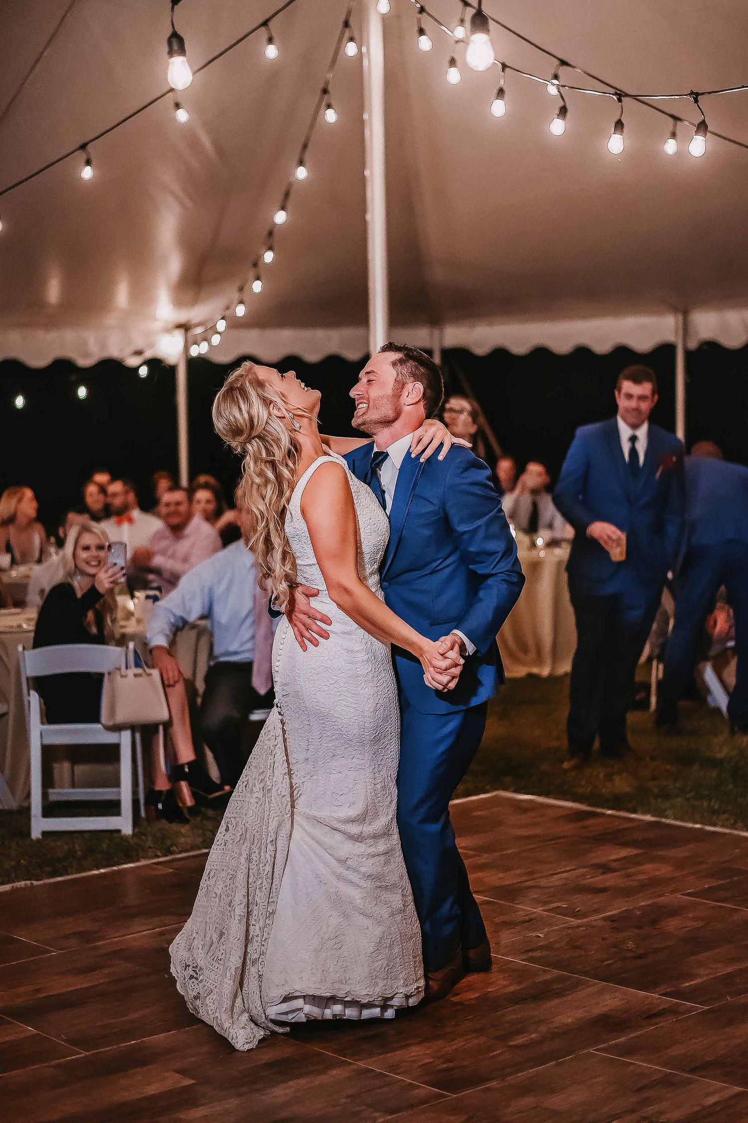 Bride and groom share their first dance under string lights in a white wedding tent with guests watching.