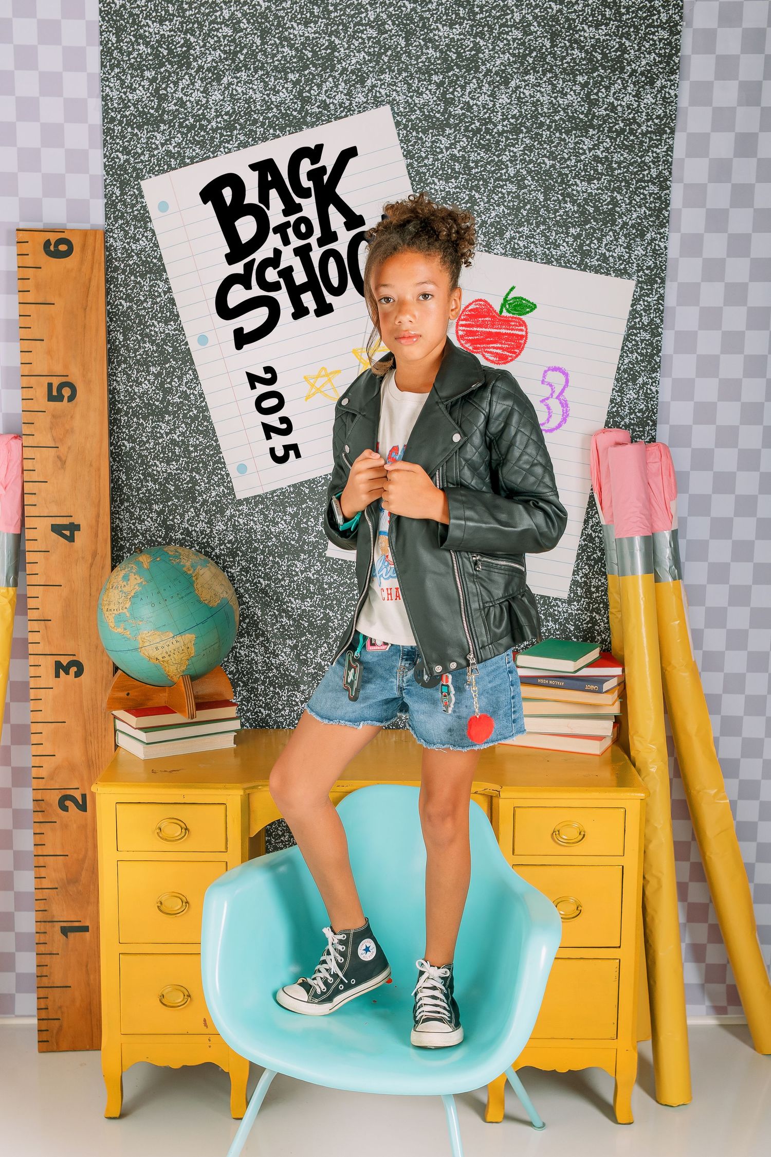 Back to school photo setup with yellow furniture, teal chair, ruler props, and casual outfit with black leather jacket.