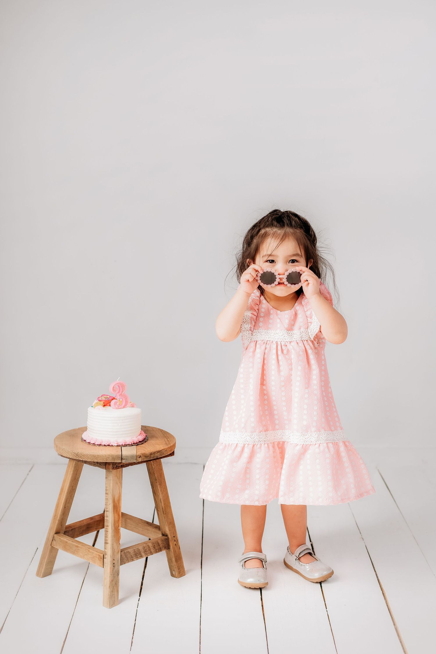 Young child in pink dress stands next to wooden stool with cake, making playful expression on light backdrop.