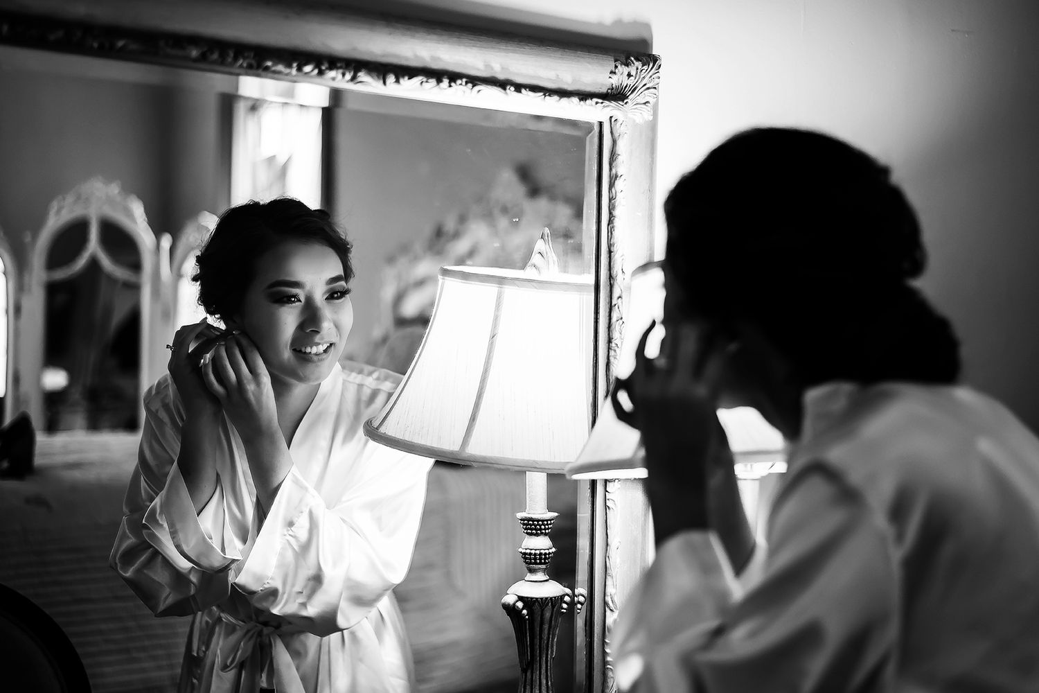 Bride in white robe smiling at mirror while applying jewelry in elegant black and white wedding preparation photo.
