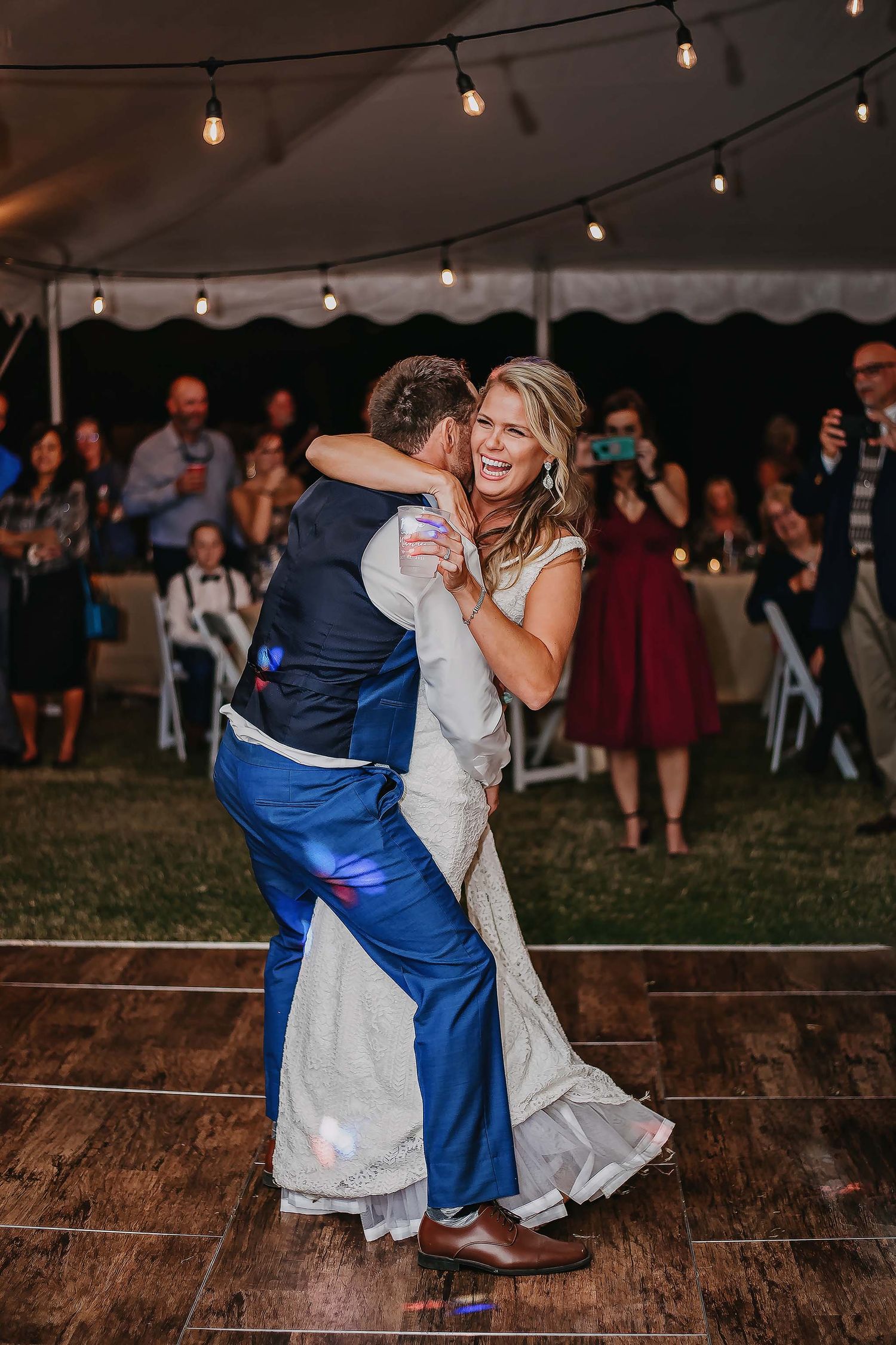 Bride laughing joyfully as groom dips her during first dance at outdoor tent wedding reception with string lights.