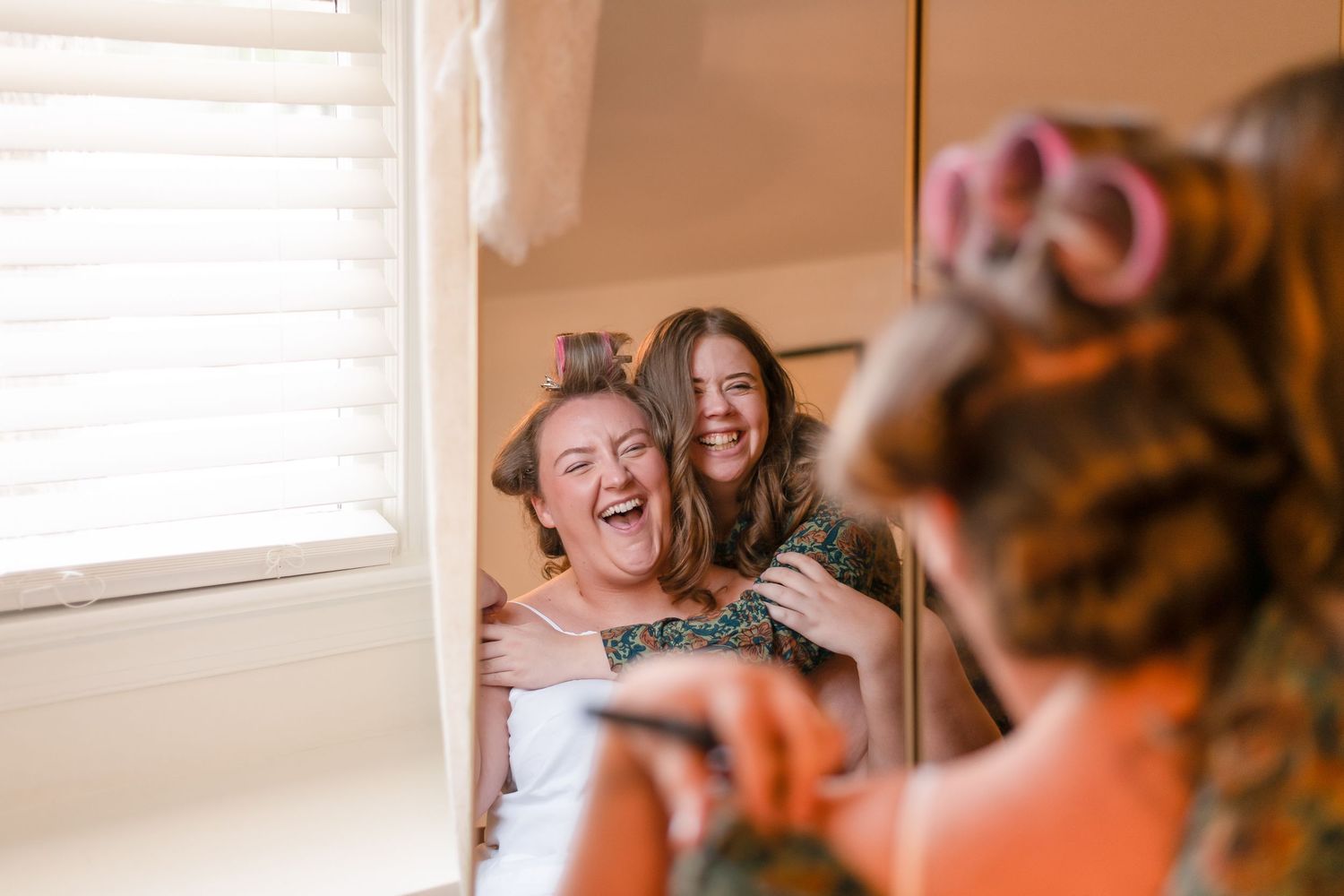 Two bridesmaids laughing joyfully with hair rollers during wedding preparations in a bright room.