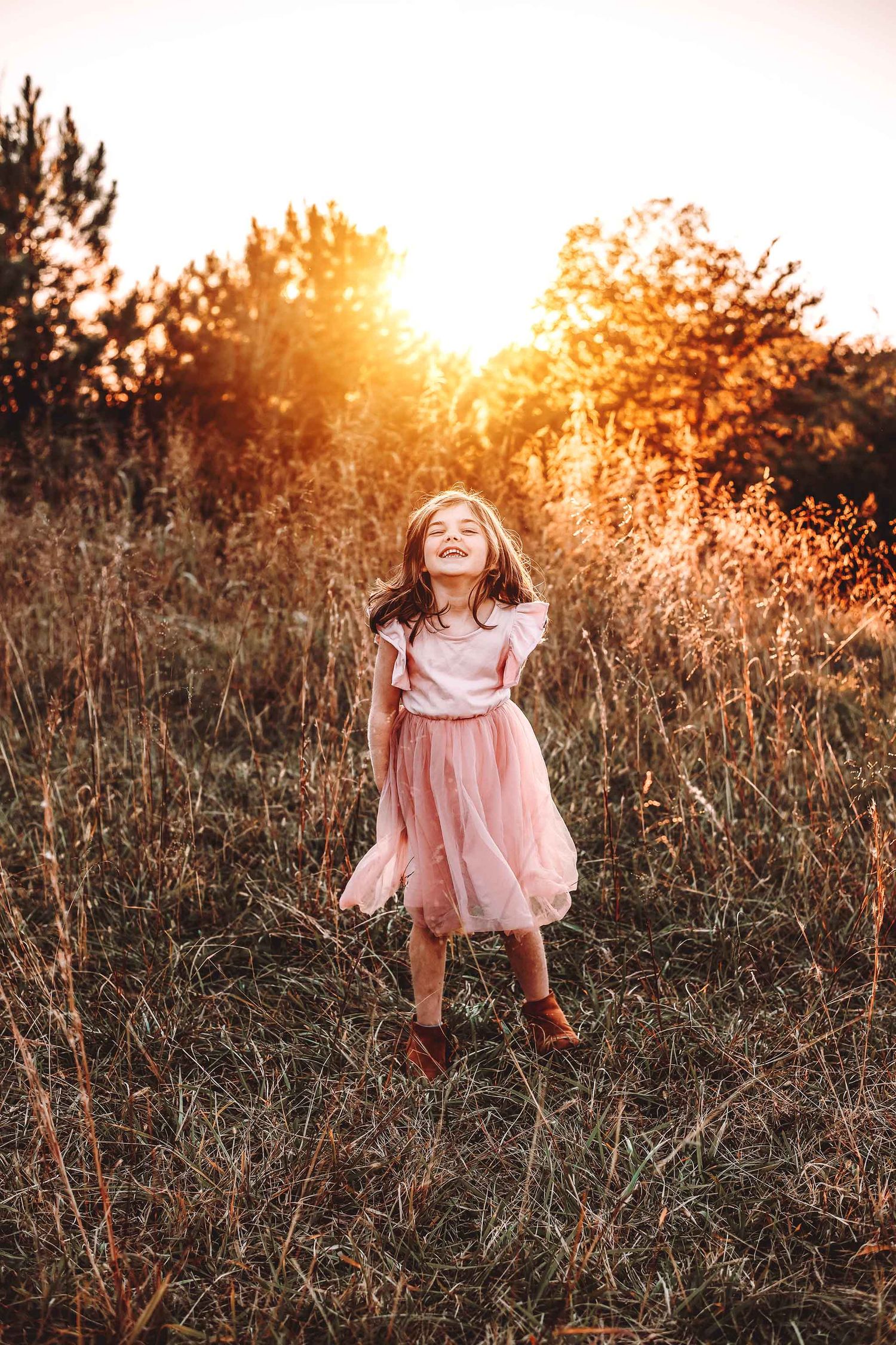 A joyful little girl in a pink tulle dress laughs in a golden sunset field, surrounded by tall grass and trees.