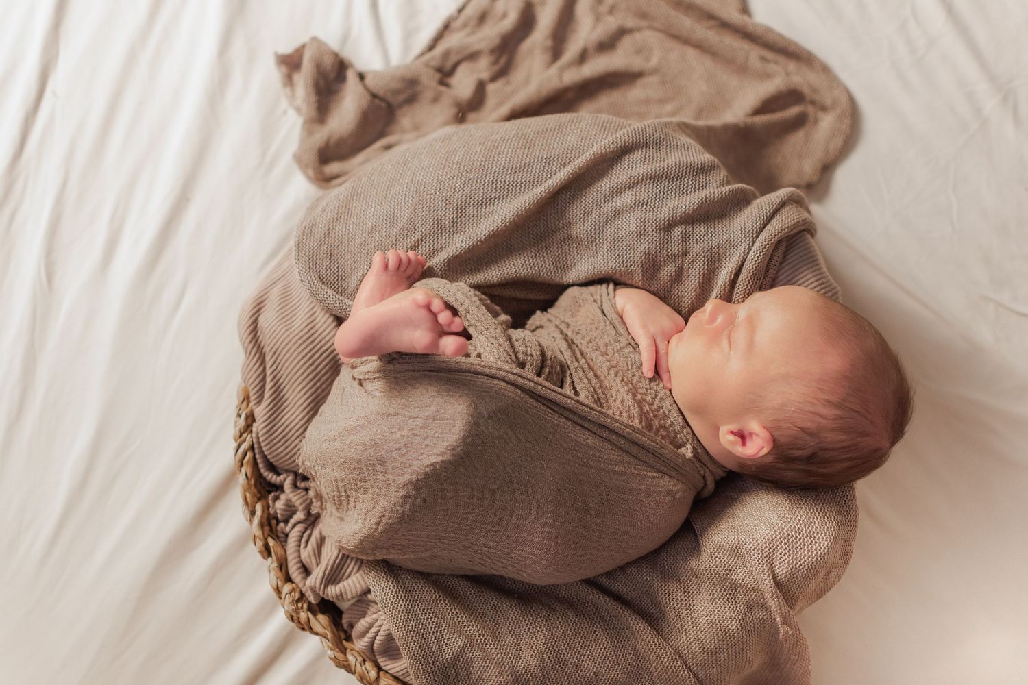 Sleeping newborn baby wrapped in a taupe knit blanket nestled in a wicker basket on white bedding.