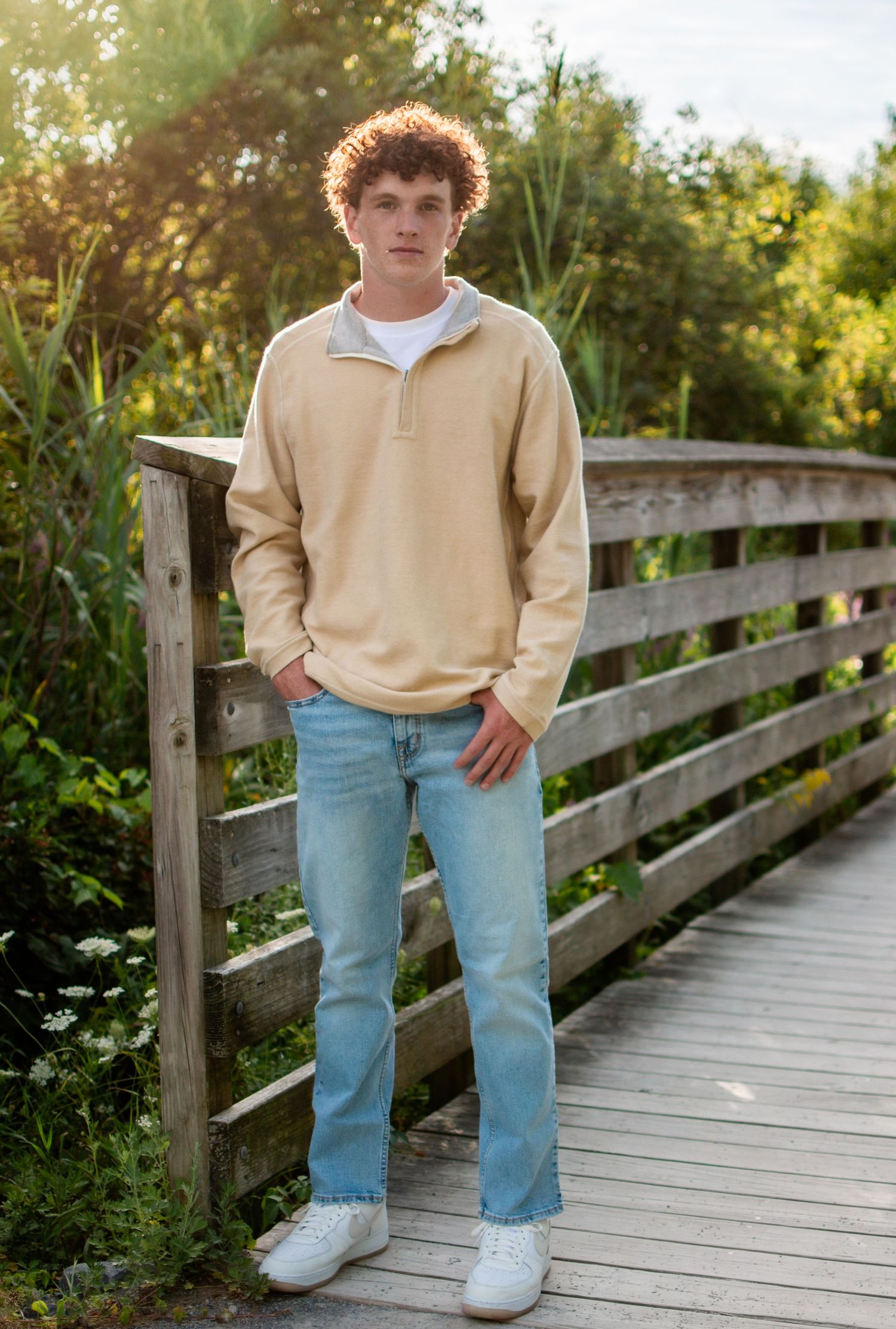 Teen boy in tan pullover and jeans standing on wooden bridge surrounded by nature.
