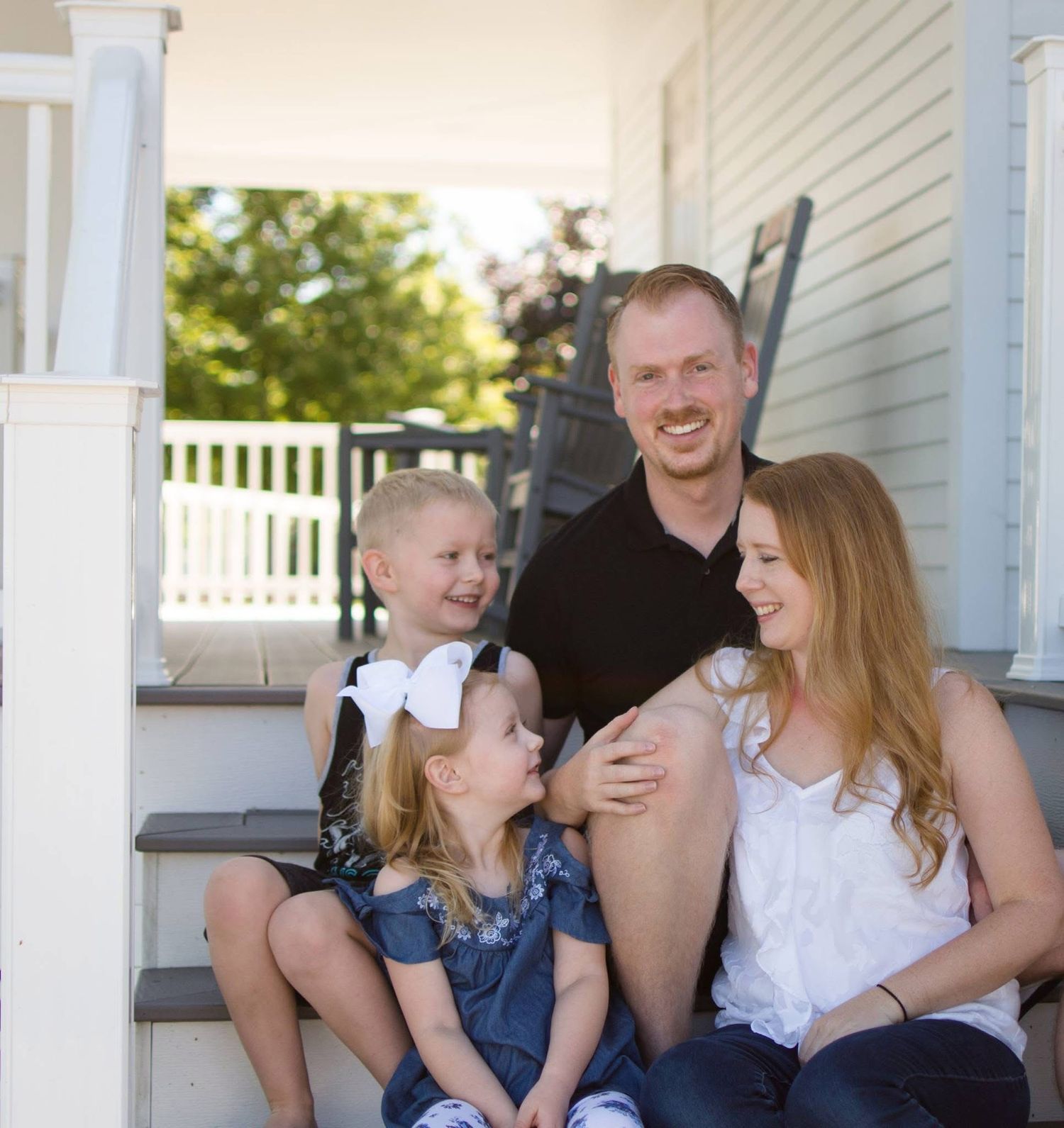 Happy family of four sitting on porch steps, dad in black shirt with two young daughters and redhead mom.
