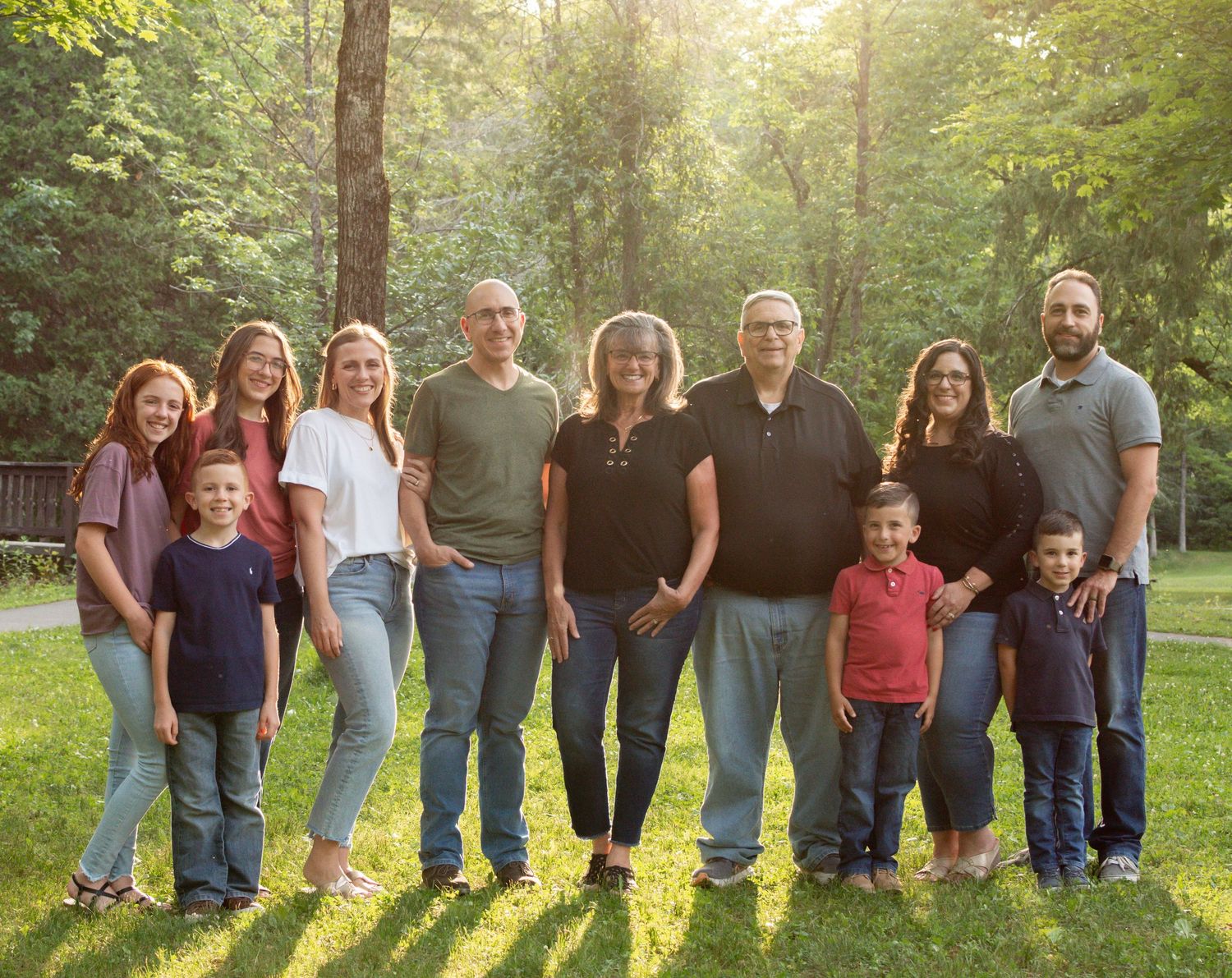 Large family group smiling together outdoors in a sunlit green park setting.