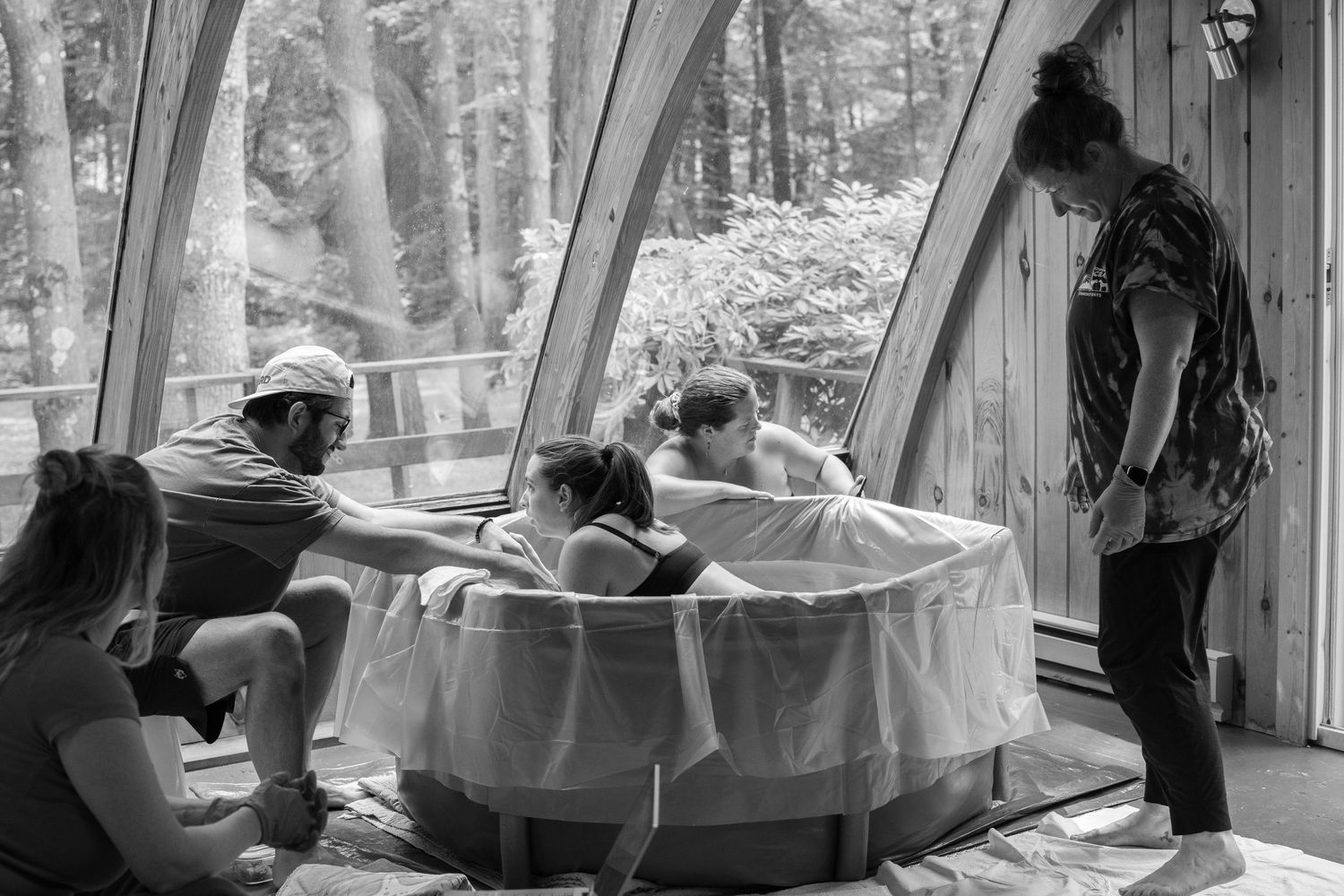 Black and white photo of a water birth with a woman in a birthing pool surrounded by supportive attendants indoors.