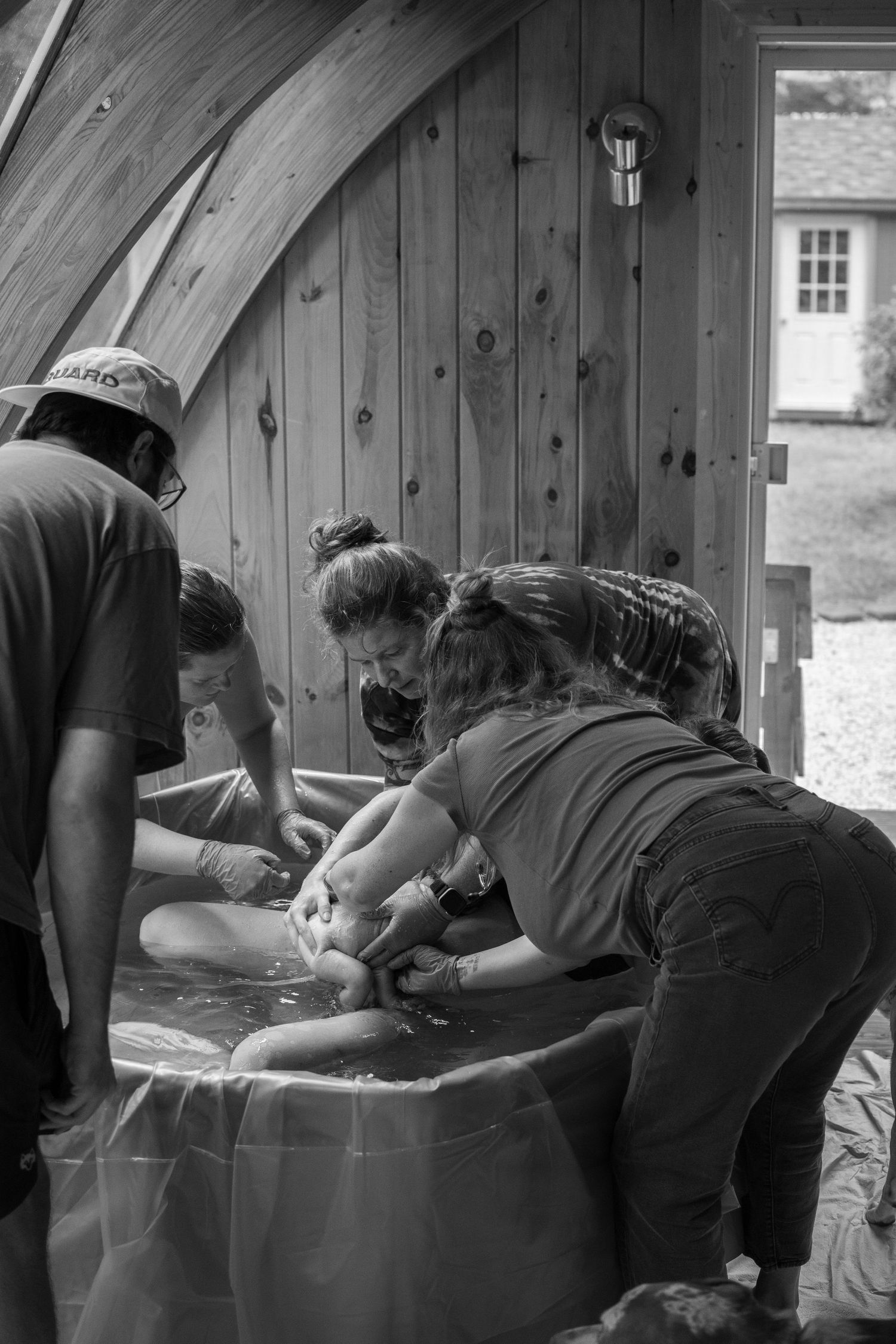Black and white photo of three people leaning over a water birth tub in a wooden cabin setting.