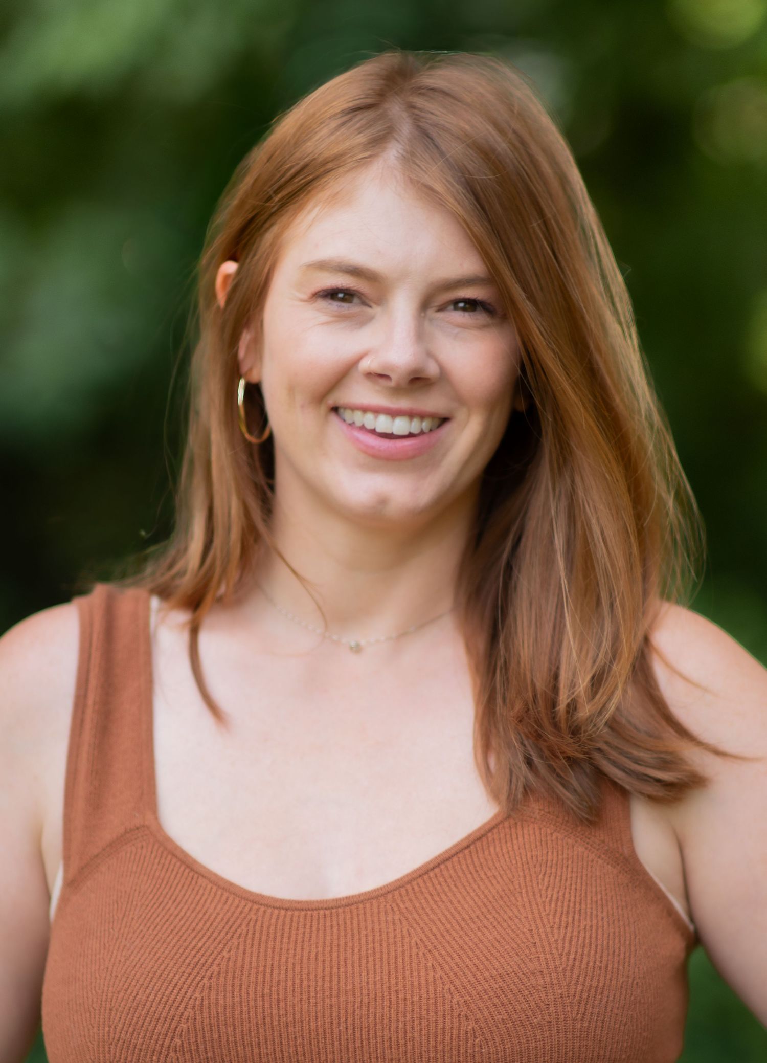 Smiling redhead woman in a rust-colored tank top poses outdoors with green bokeh background.