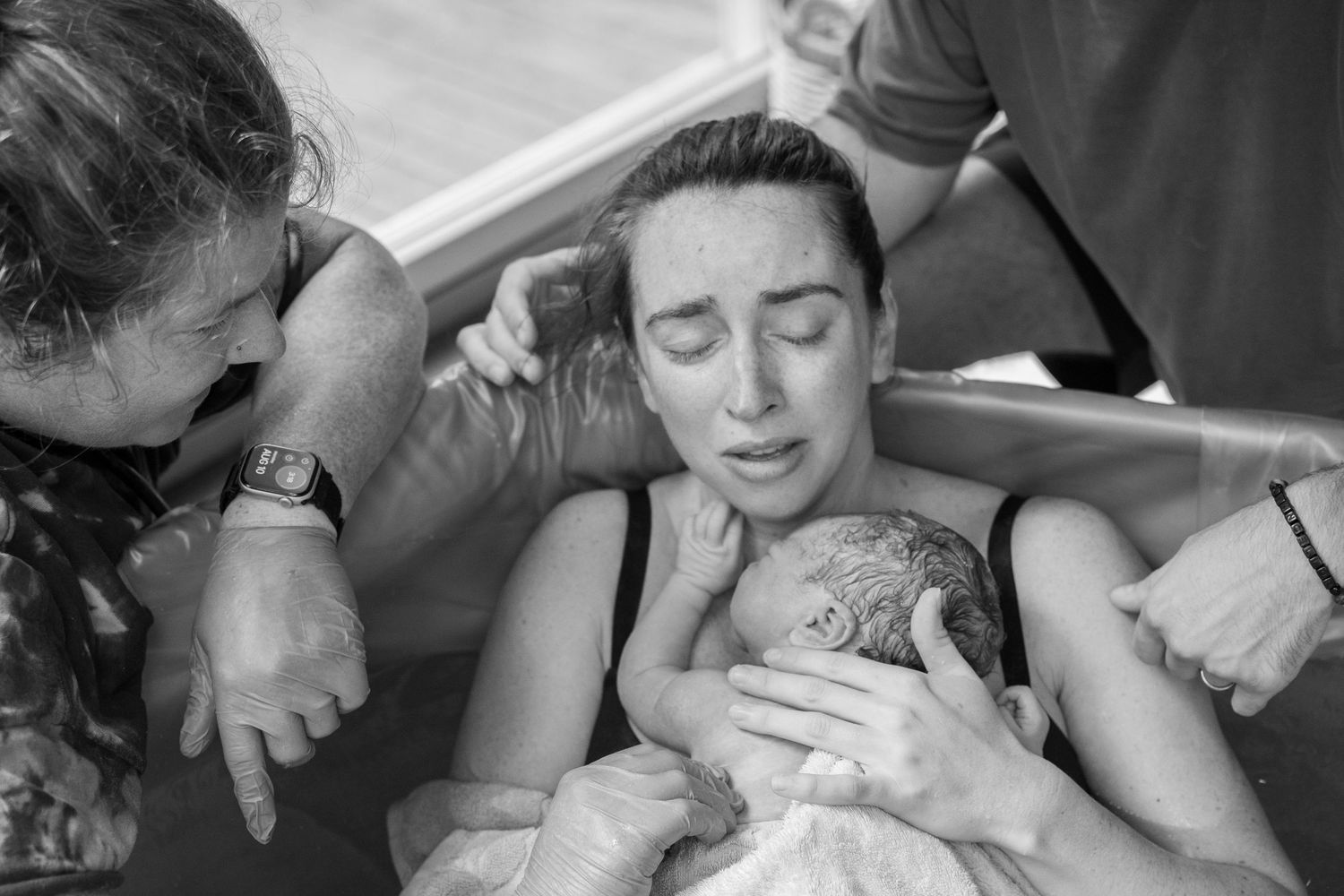 Black and white photo of a woman holding her newborn baby during a water birth, supported by caregivers.