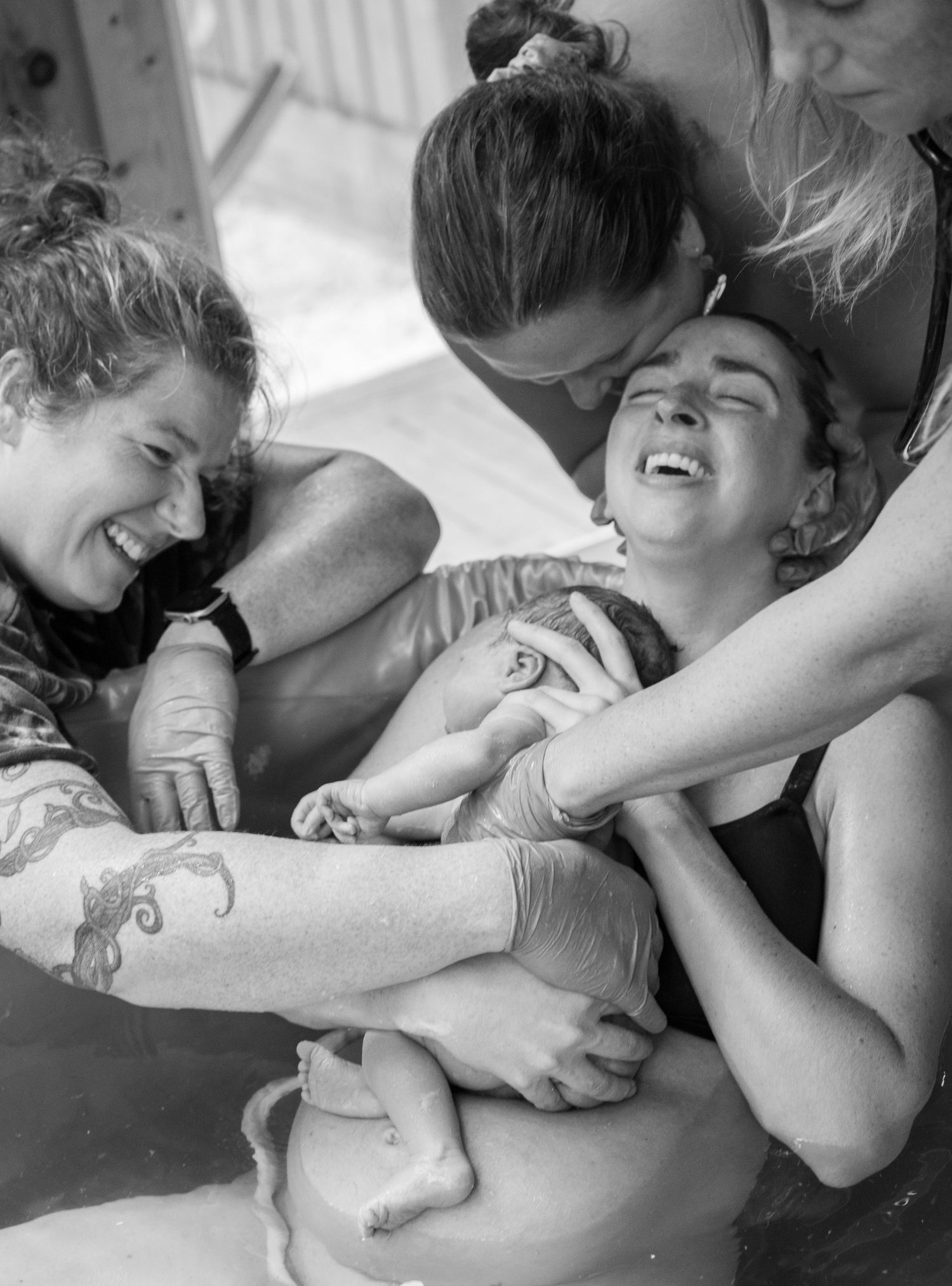 Black and white photo of a joyful water birth, with a newborn baby being held by smiling women in a birthing pool.