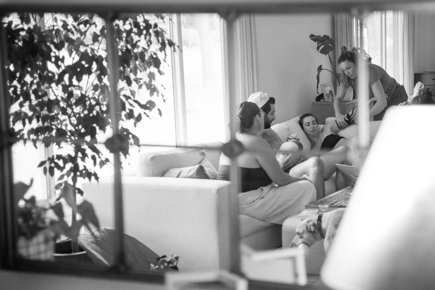 Black and white photo of people relaxing on a white sofa in a bright living room with large windows and plants.
