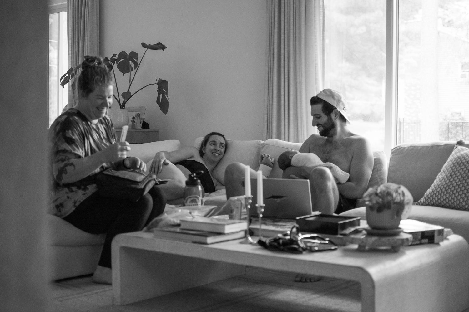 Three friends laughing and relaxing together in a bright modern living room, captured in black and white photography. Home birth postpartum.