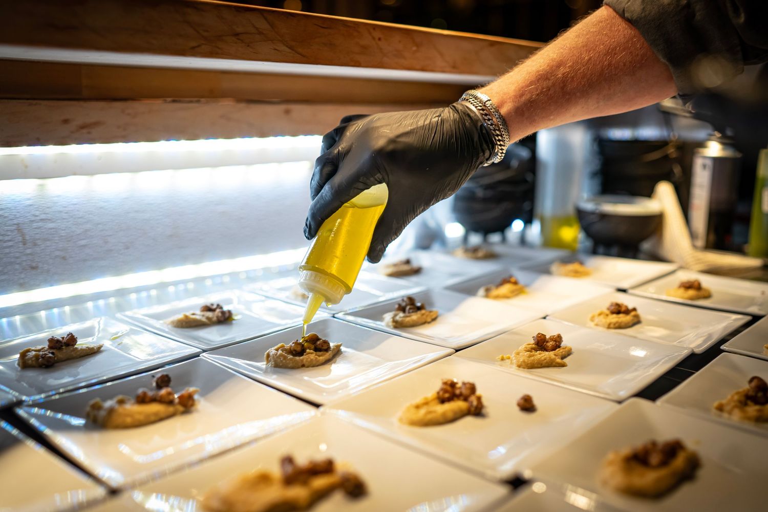 A person wearing a black glove drizzles yellow sauce onto small square plates of hummus topped with chickpeas, arranged neatly in rows.