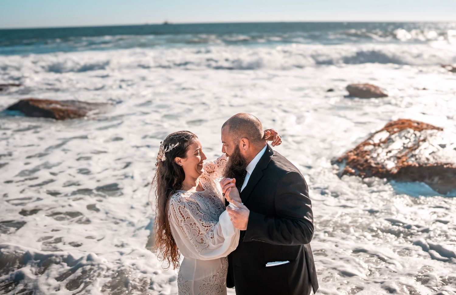A couple dressed in formal attire stand close, smiling at each other on a rocky beach. The woman wears a white dress, and the man is in a da